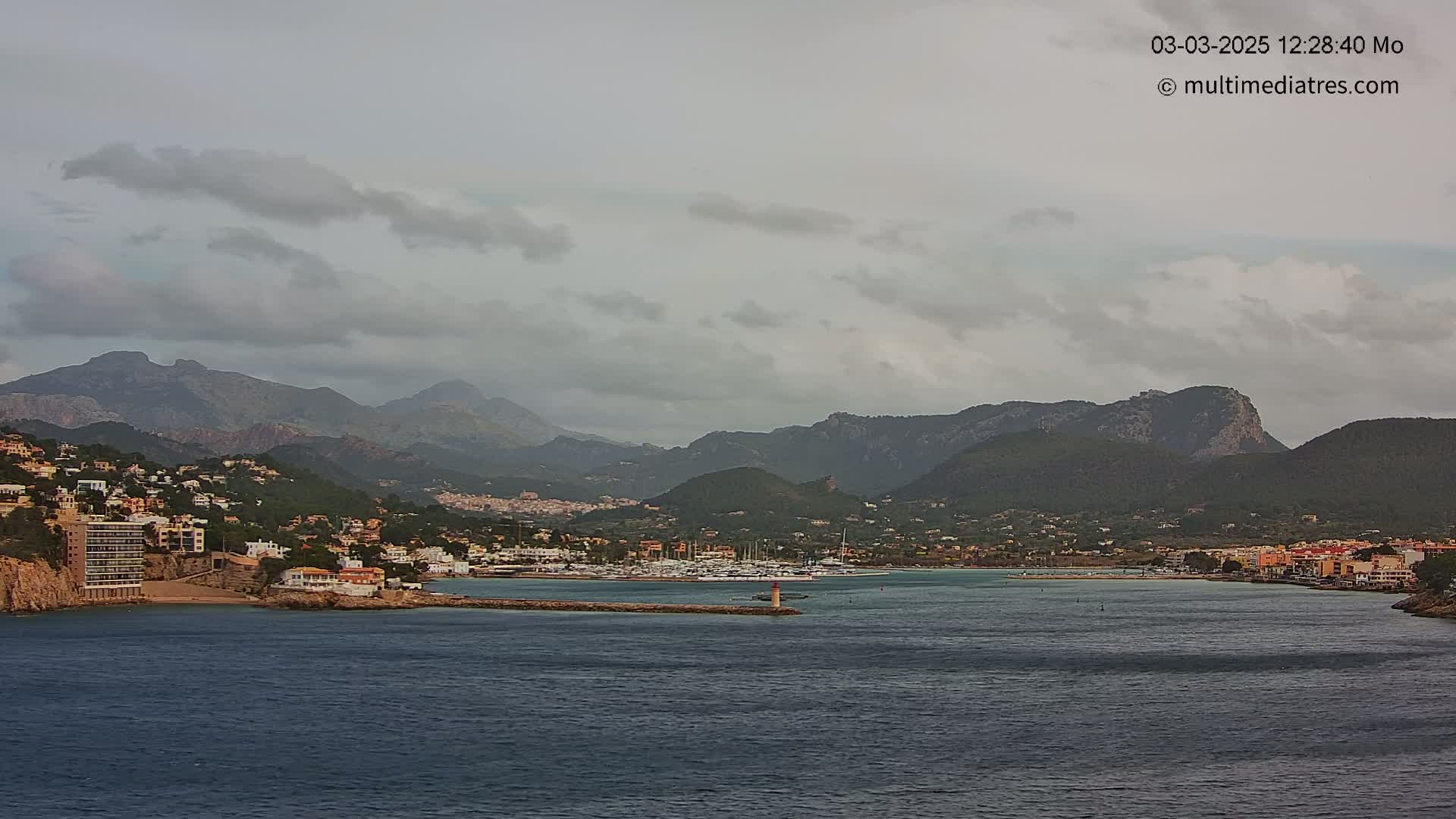 A coastal town nestled at the foot of mountains under a cloudy sky, with a calm body of water in the foreground and a marina visible in the middle ground.