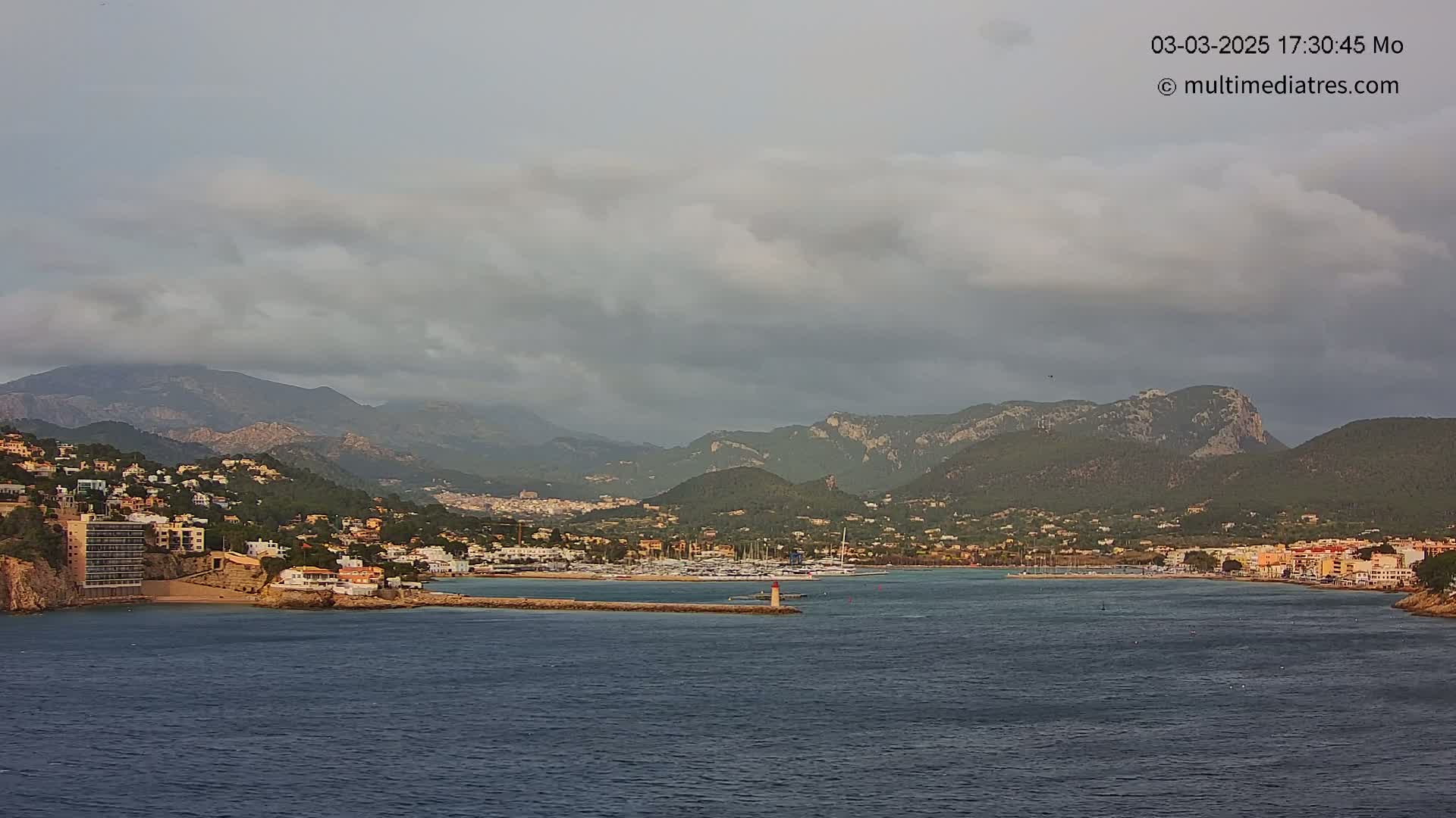 A coastal town nestled against a mountain range under a cloudy sky sits on a body of dark water with a marina visible.
