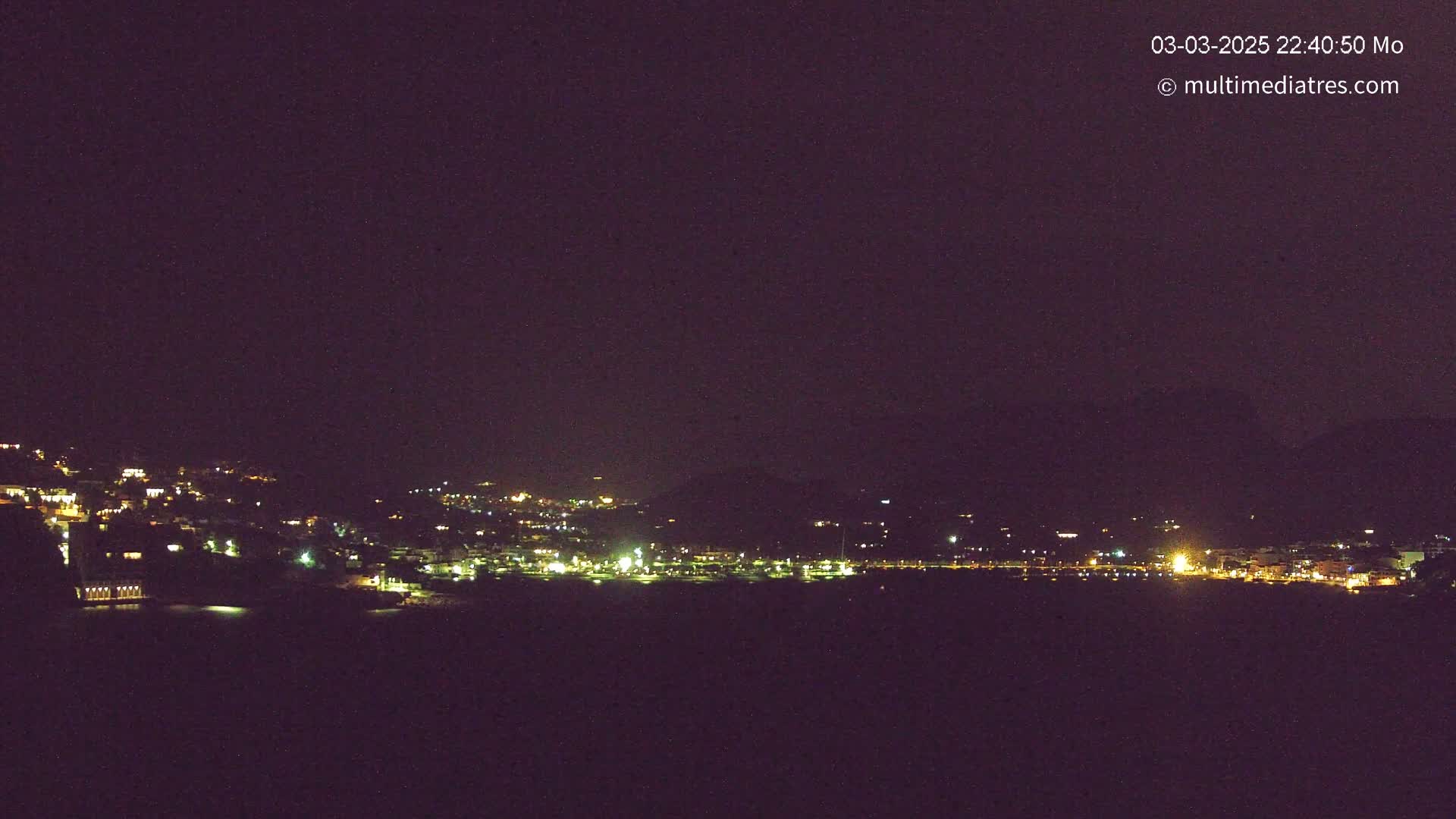 A nighttime long shot of a town nestled on a hillside, illuminated by numerous scattered lights under a clear, dark sky.