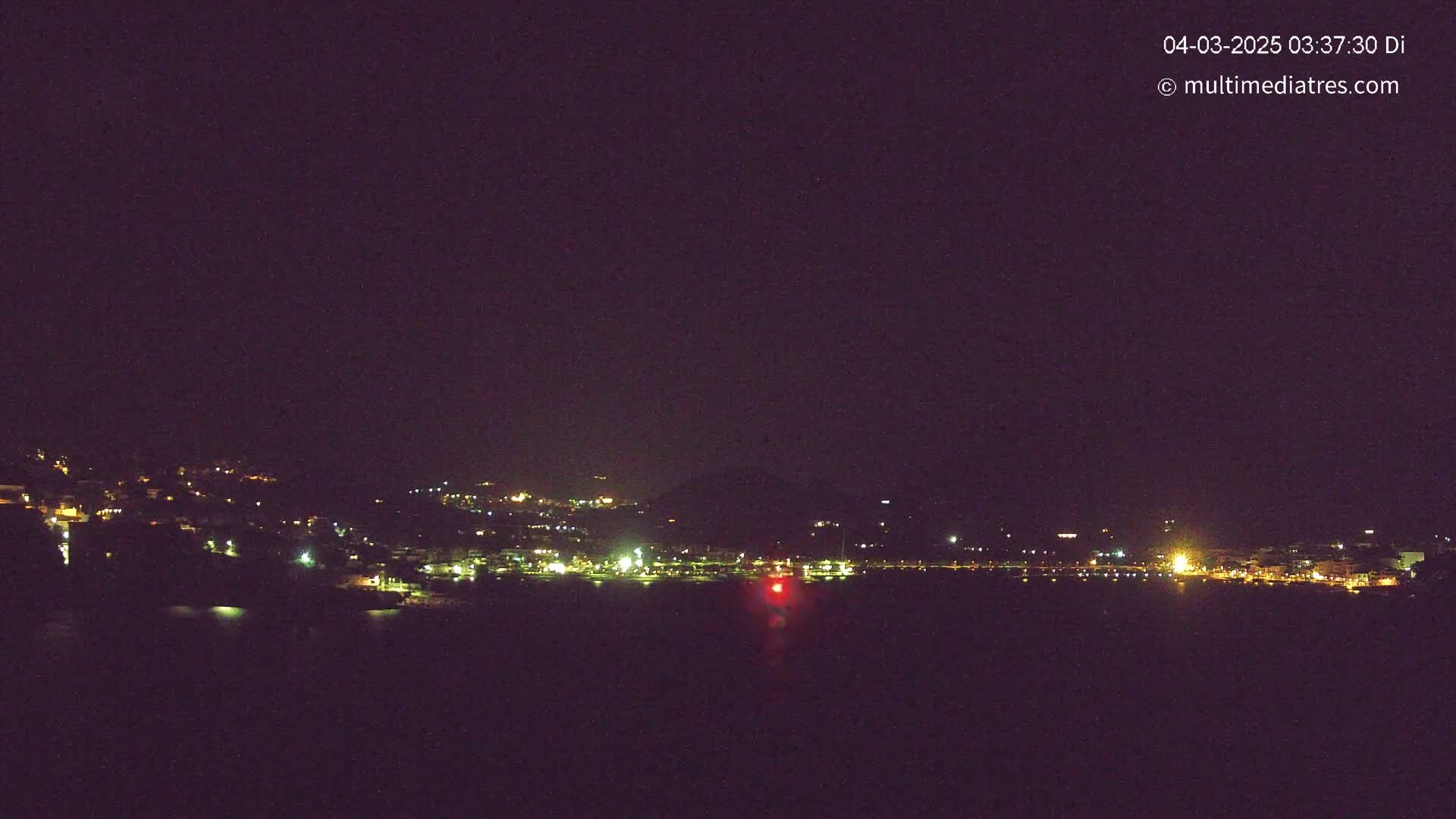 A nighttime view of a coastal town with scattered lights across a hillside and along a waterfront, under a clear, dark sky.