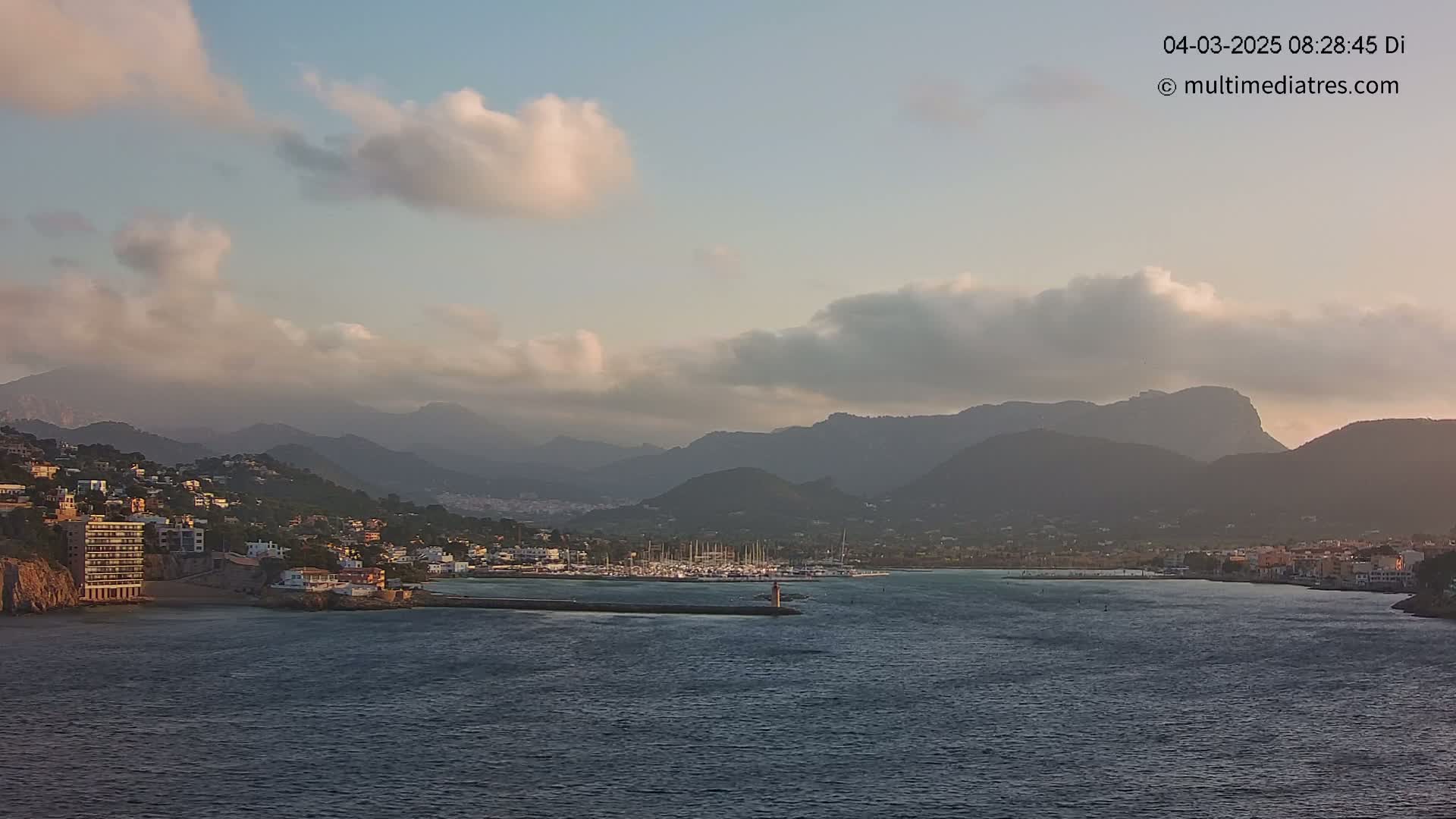 A partly cloudy sunrise illuminates a coastal town nestled between mountains and a calm body of water with a marina filled with sailboats.