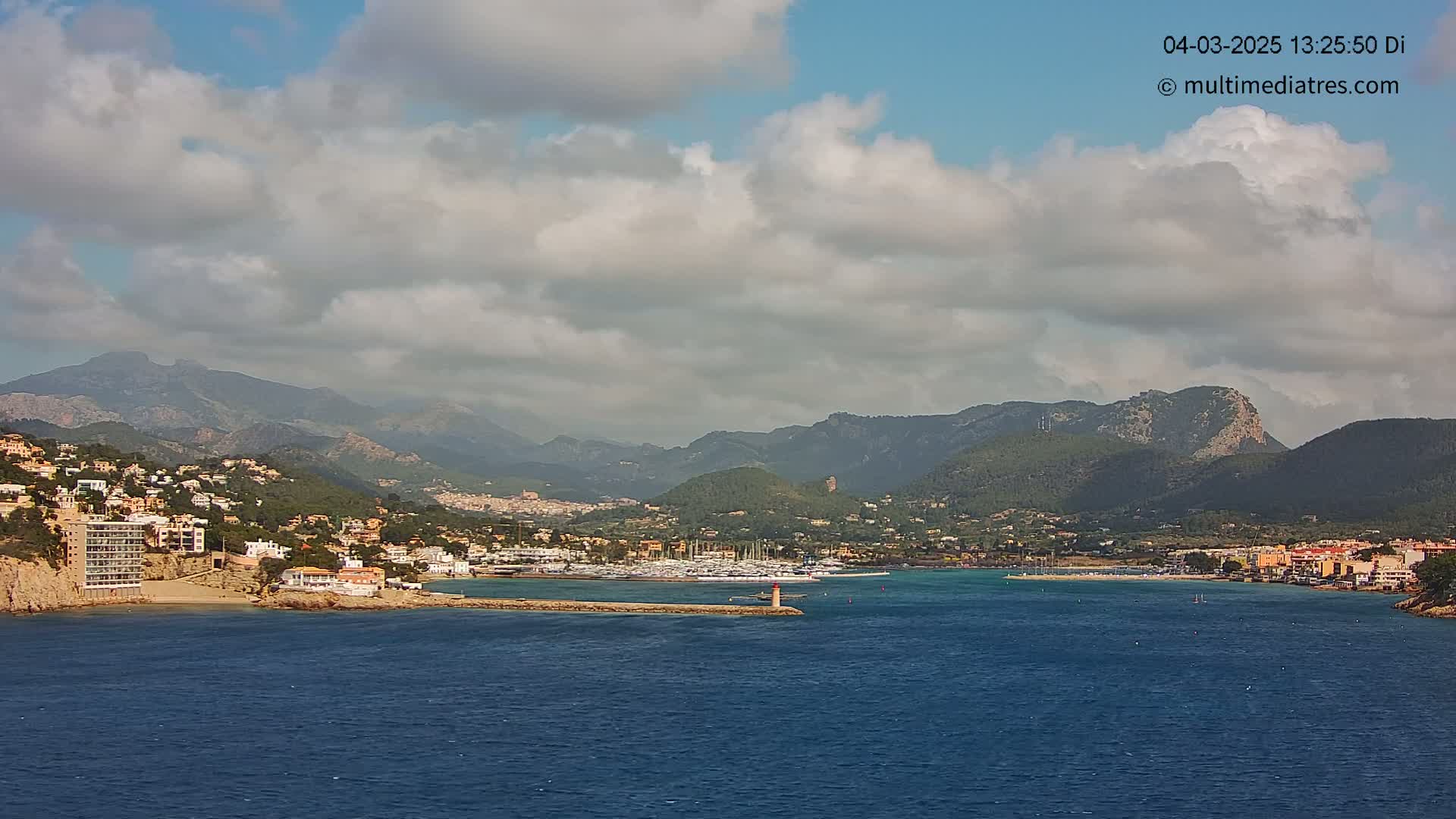 A partly cloudy sky overlooks a coastal town nestled between mountains and a calm blue sea, with a marina filled with boats.