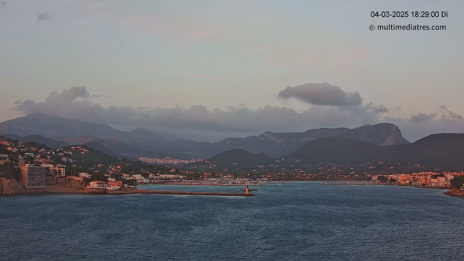 A coastal town nestled against mountains under a partly cloudy twilight sky sits on a calm body of water with a small lighthouse visible in the harbor.