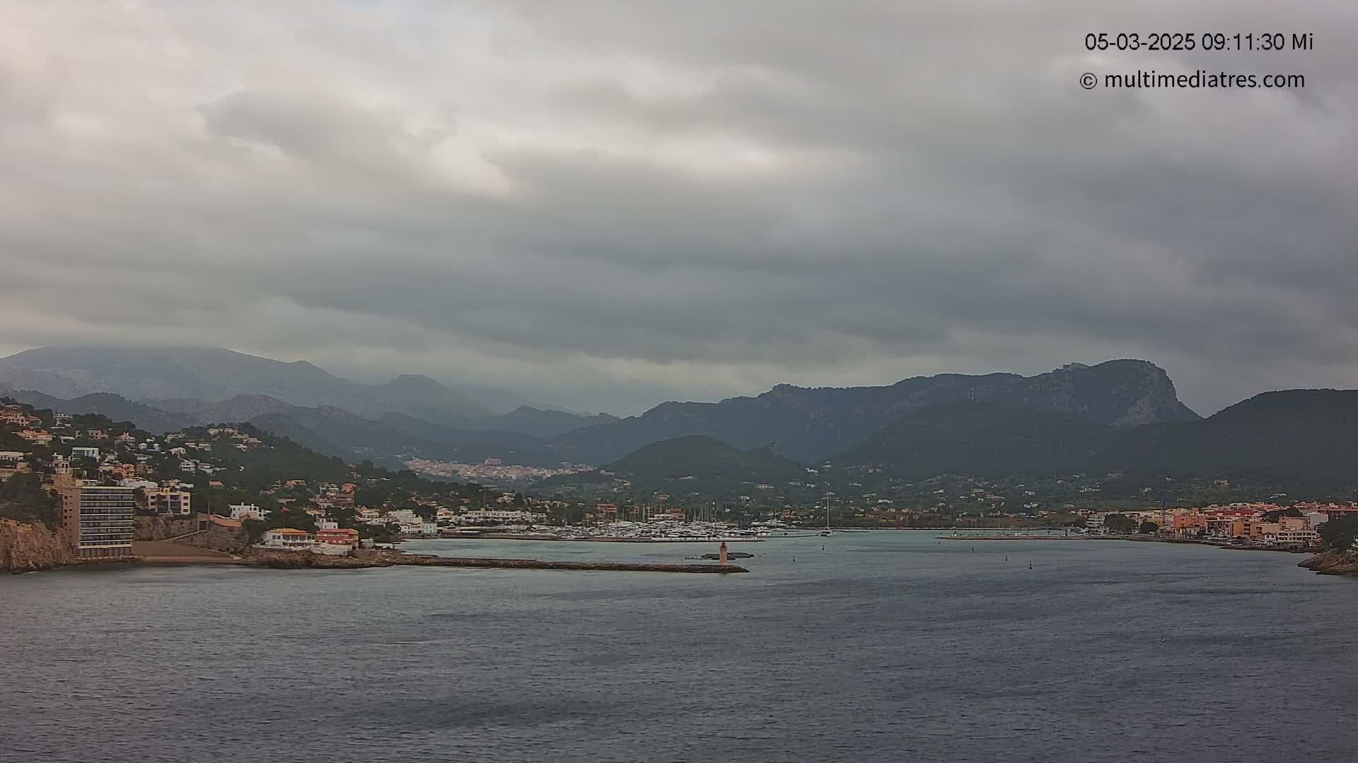 A coastal town nestled against mountains under an overcast sky, with a calm harbor filled with boats.