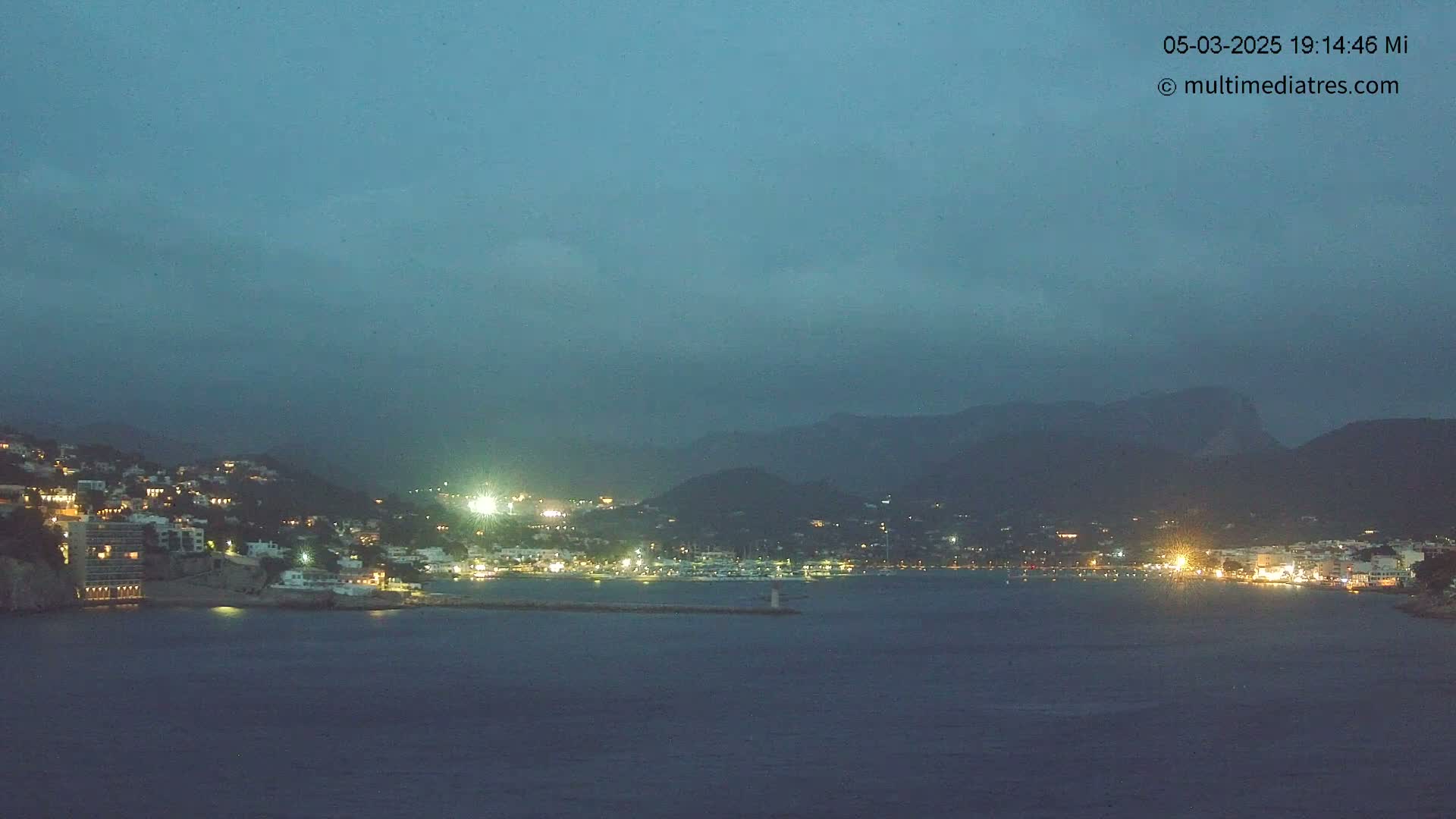 A nighttime view of a coastal town illuminated with lights, nestled between dark mountains under a dusky sky.