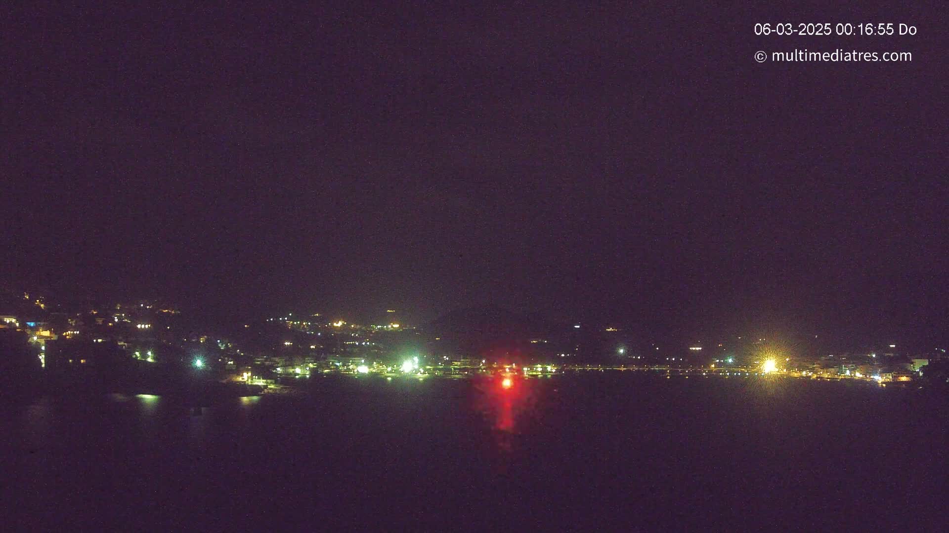 A nighttime view of a town situated on a hillside overlooking a body of water, with scattered lights illuminating the scene.