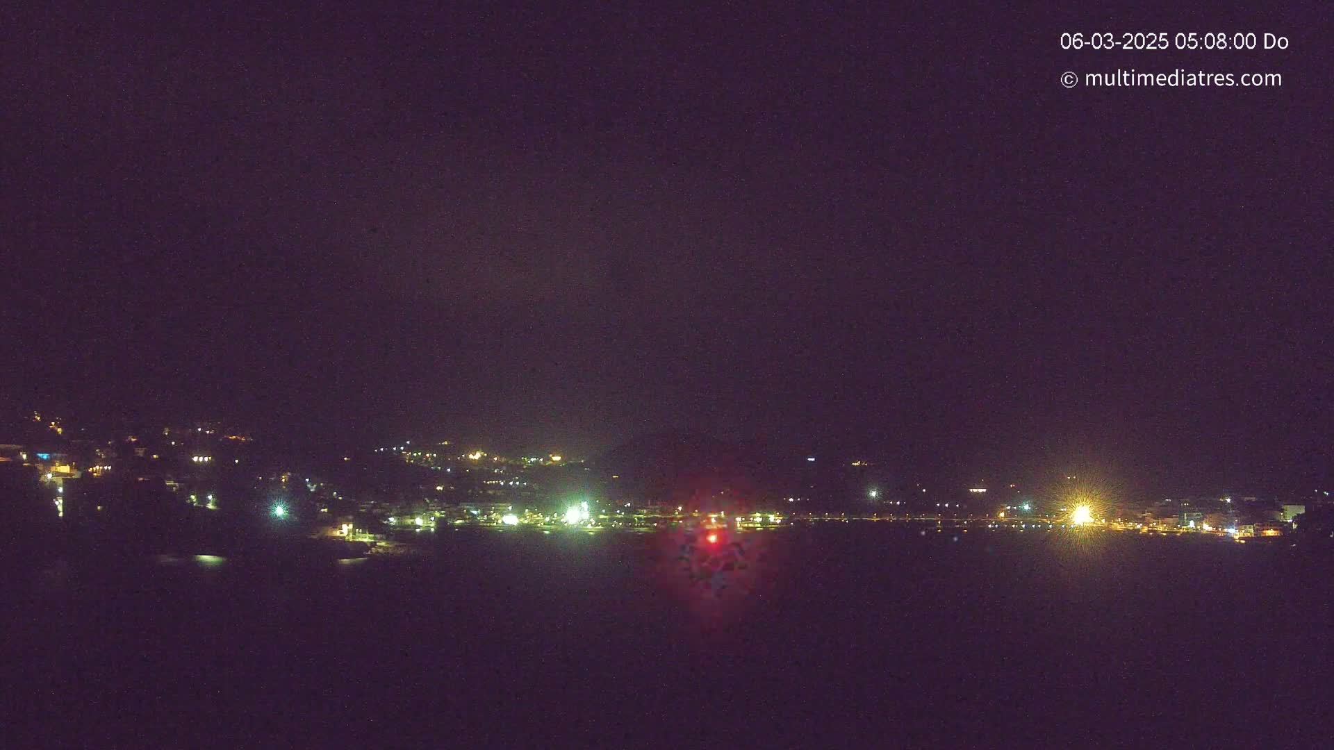 A nighttime long shot shows a town's lights spread across a hillside and reflected in dark water under a clear, dark sky.