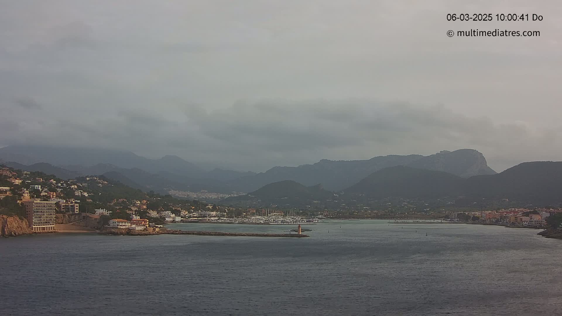 A coastal town nestled between mountains under an overcast sky is seen from across a calm body of water.