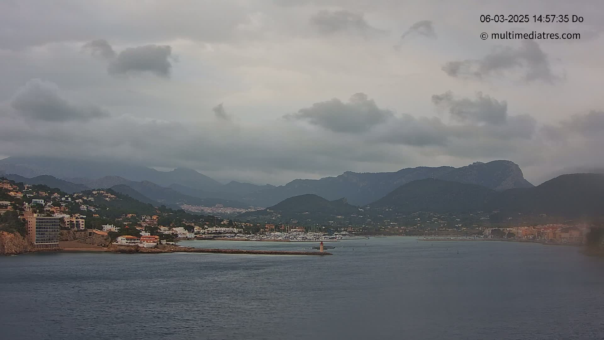 A coastal town nestled between dark mountains is viewed from across a calm, gray body of water under an overcast sky.