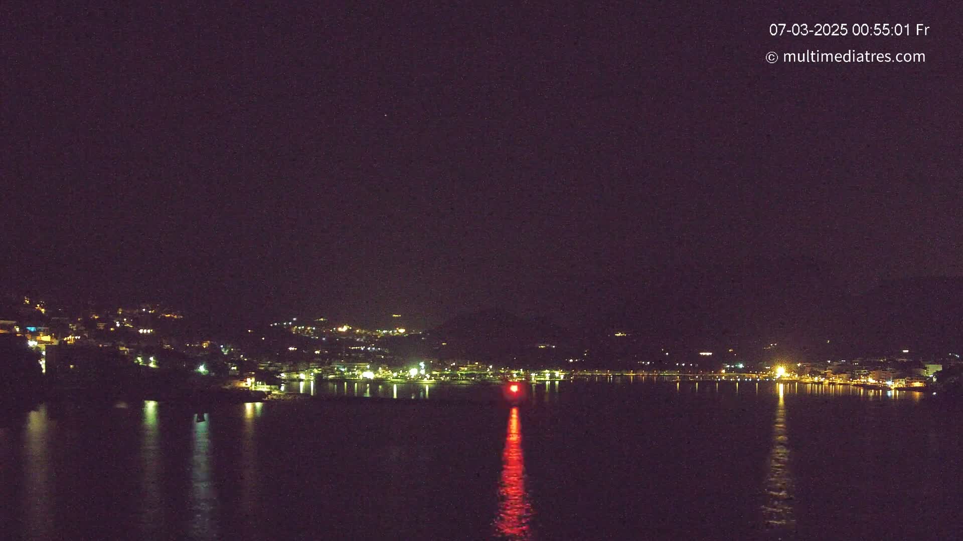 A nighttime view of a town's lights along a dark body of water, reflecting the lights, with a single red light in the water.