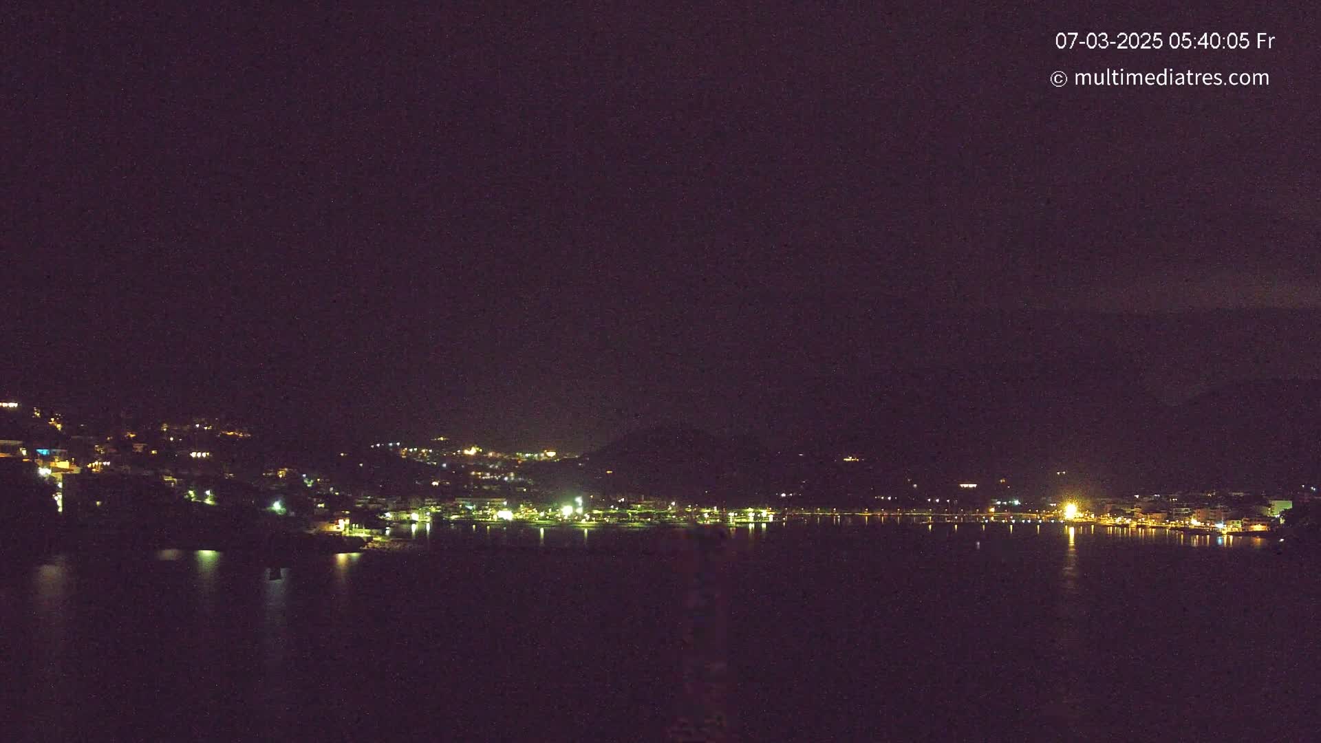 A nighttime view of a coastal town with its lights reflected in a dark body of water under a clear, starless sky.