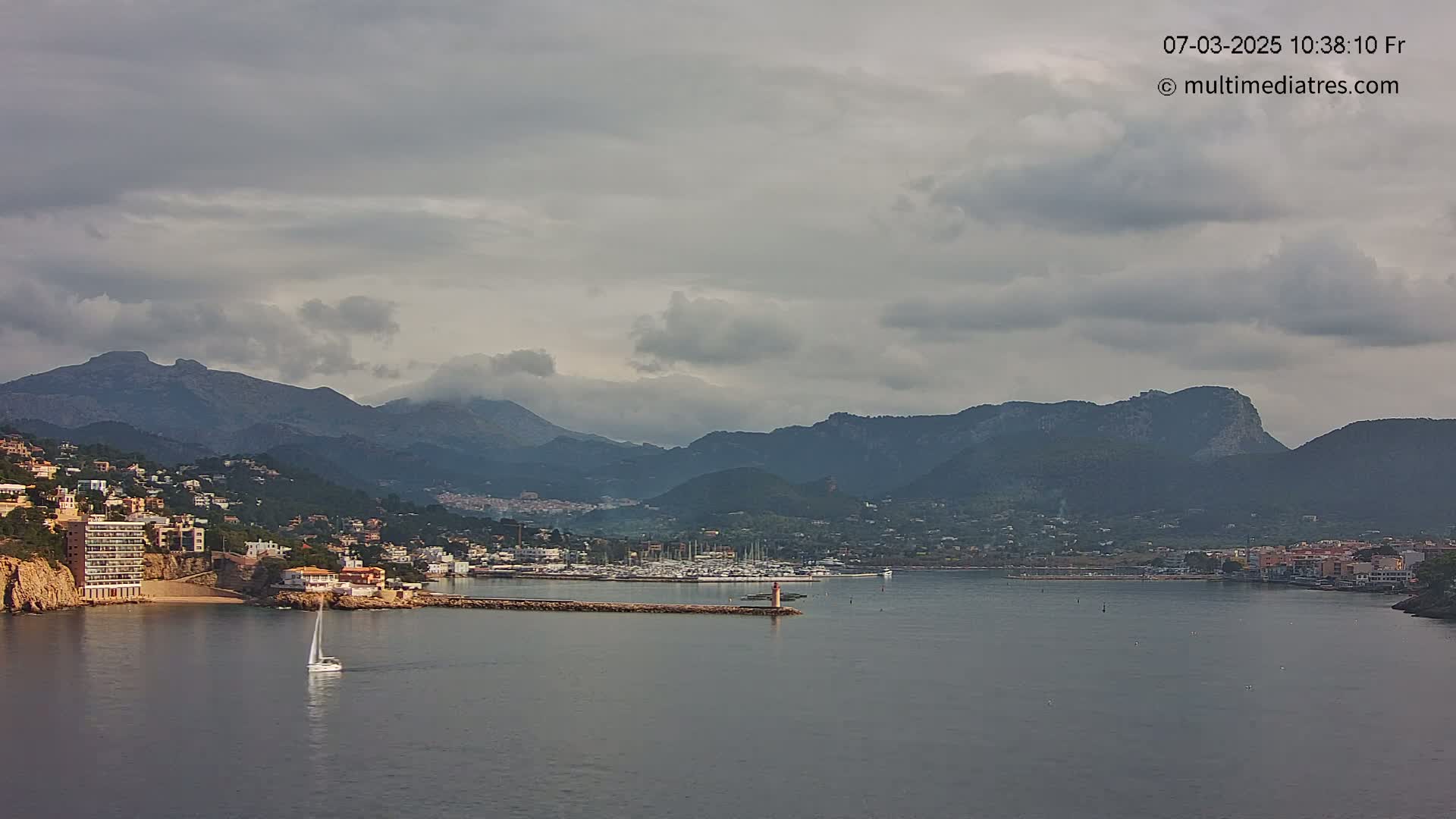 A sailboat is visible on a calm body of water, with a coastal town nestled against mountains under a cloudy sky.