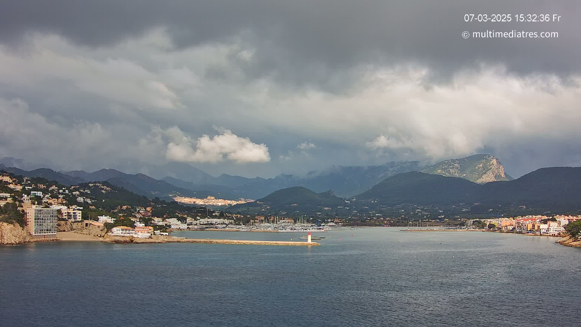 A coastal town nestled between mountains under a cloudy sky, with a harbor filled with boats.