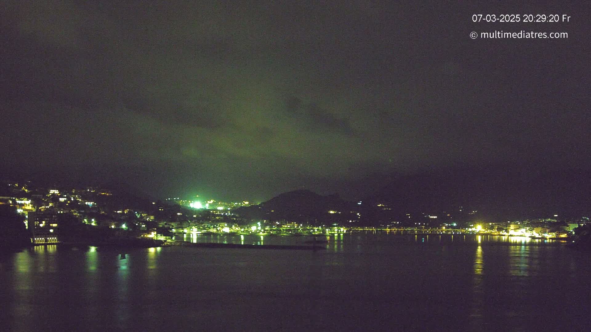A nighttime view of a town situated on a hillside overlooking a calm body of water under a dark, cloudy sky.