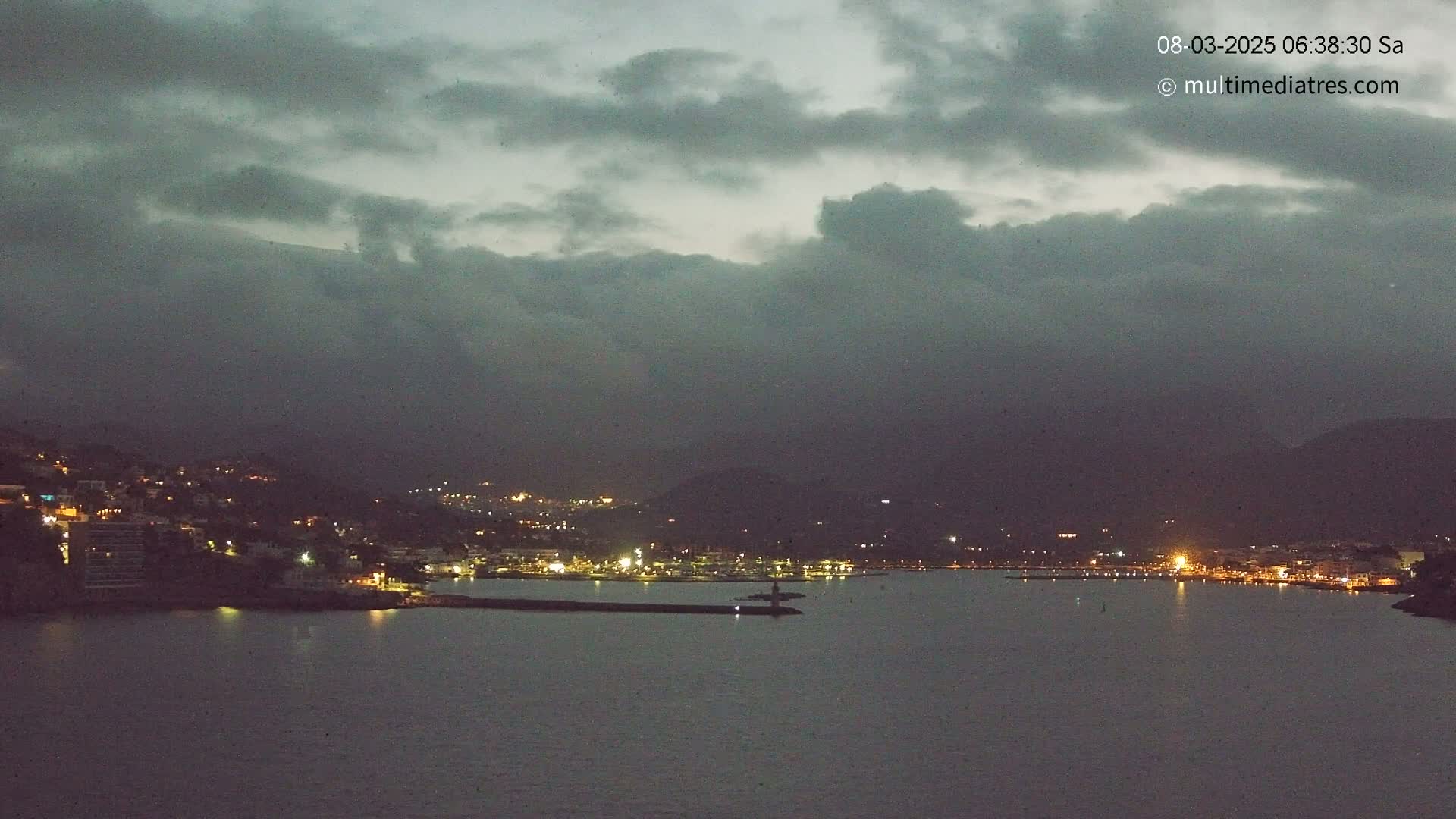 A nighttime coastal town is seen under a cloudy, dark sky, with its lights illuminating the harbor and surrounding hills.