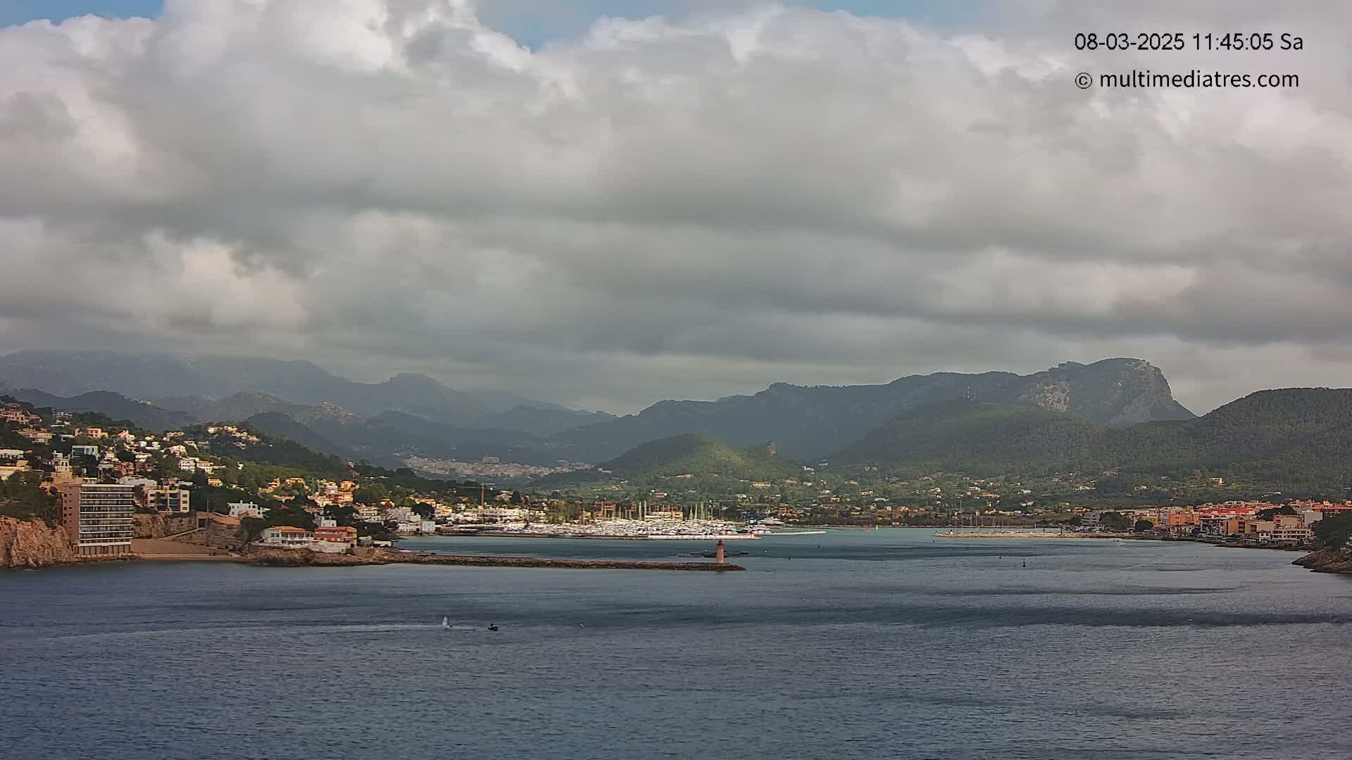 A coastal town nestled at the base of a mountain range under a cloudy sky, with a harbor filled with boats.