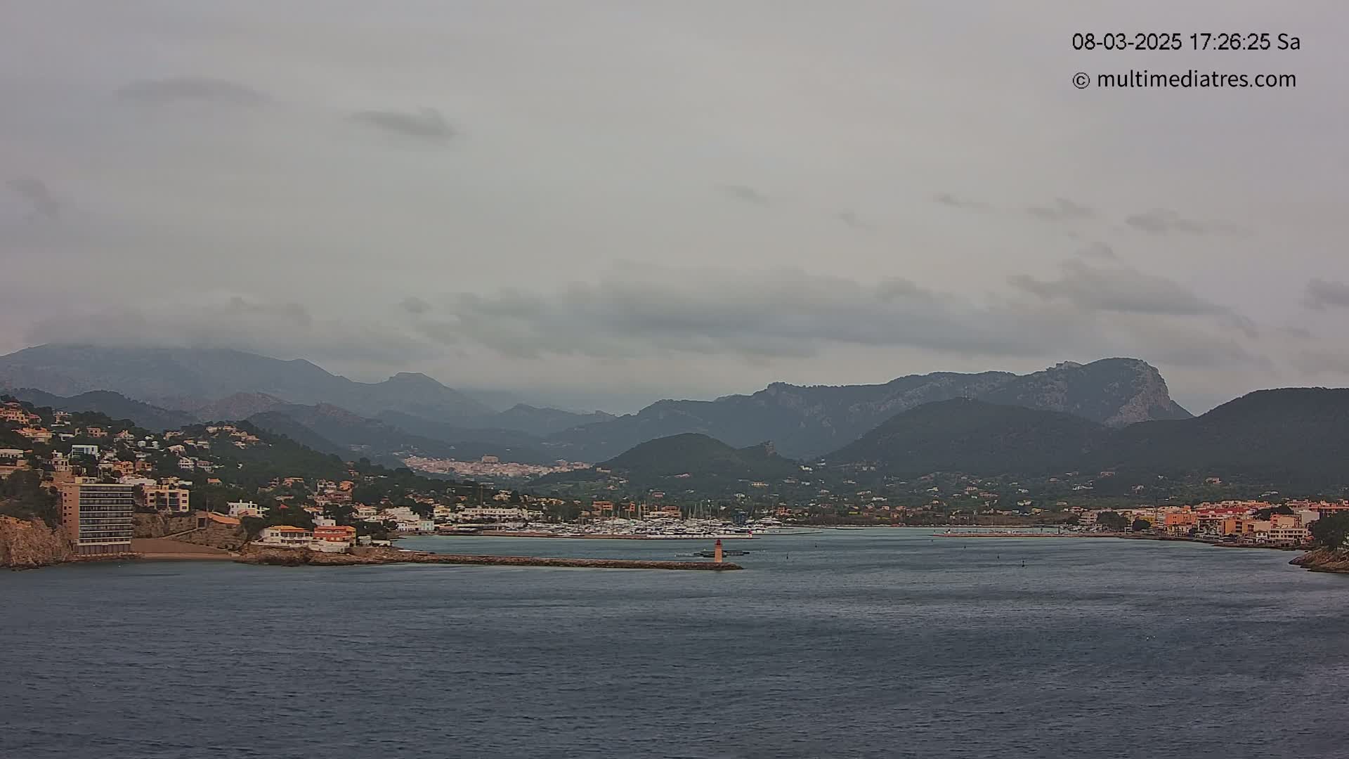 A coastal town nestled between mountains under an overcast sky is visible from across a body of water, with a small lighthouse in the foreground.