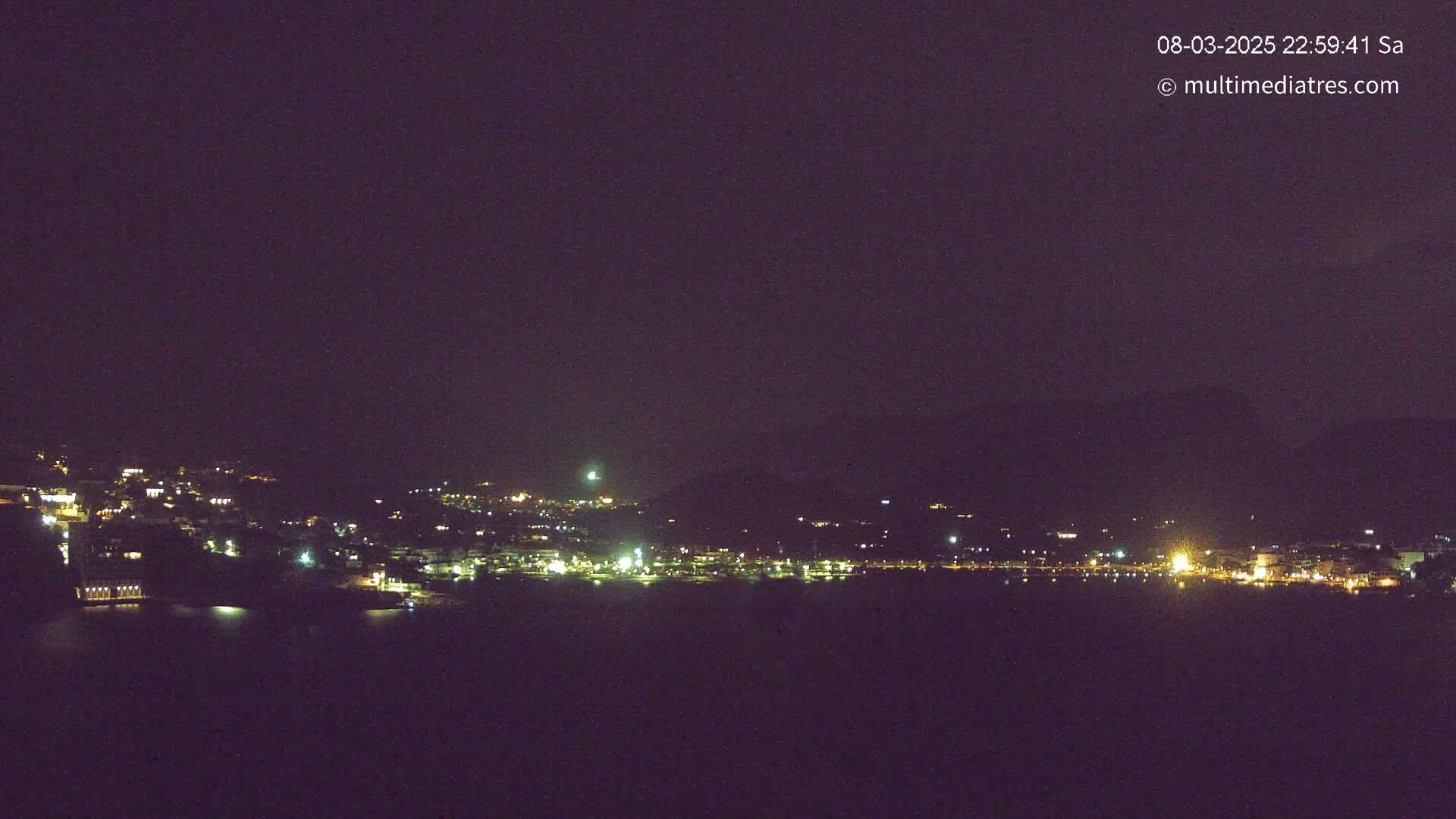 A nighttime long shot shows a town’s lights spread across a hillside next to dark water under a clear, dark sky.