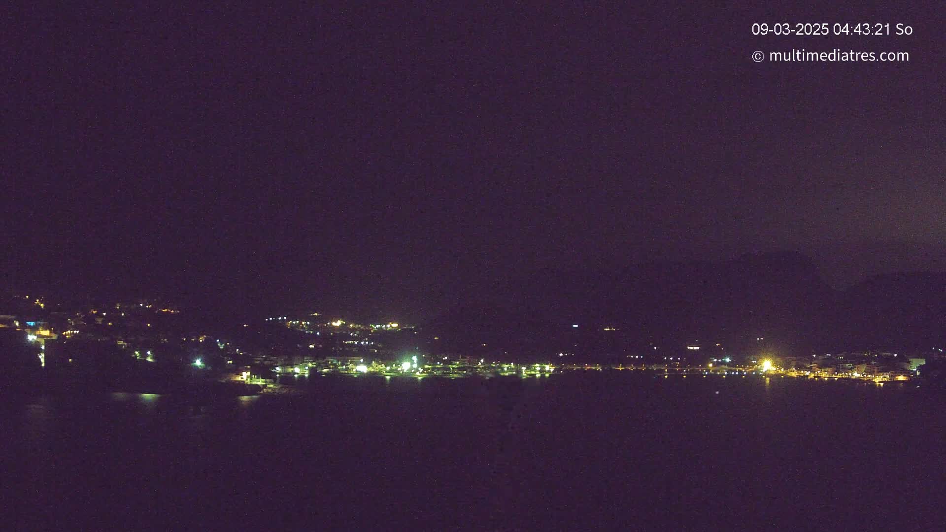 A nighttime long shot shows a dark body of water in front of a dimly lit town nestled against dark hills under a clear, dark sky.