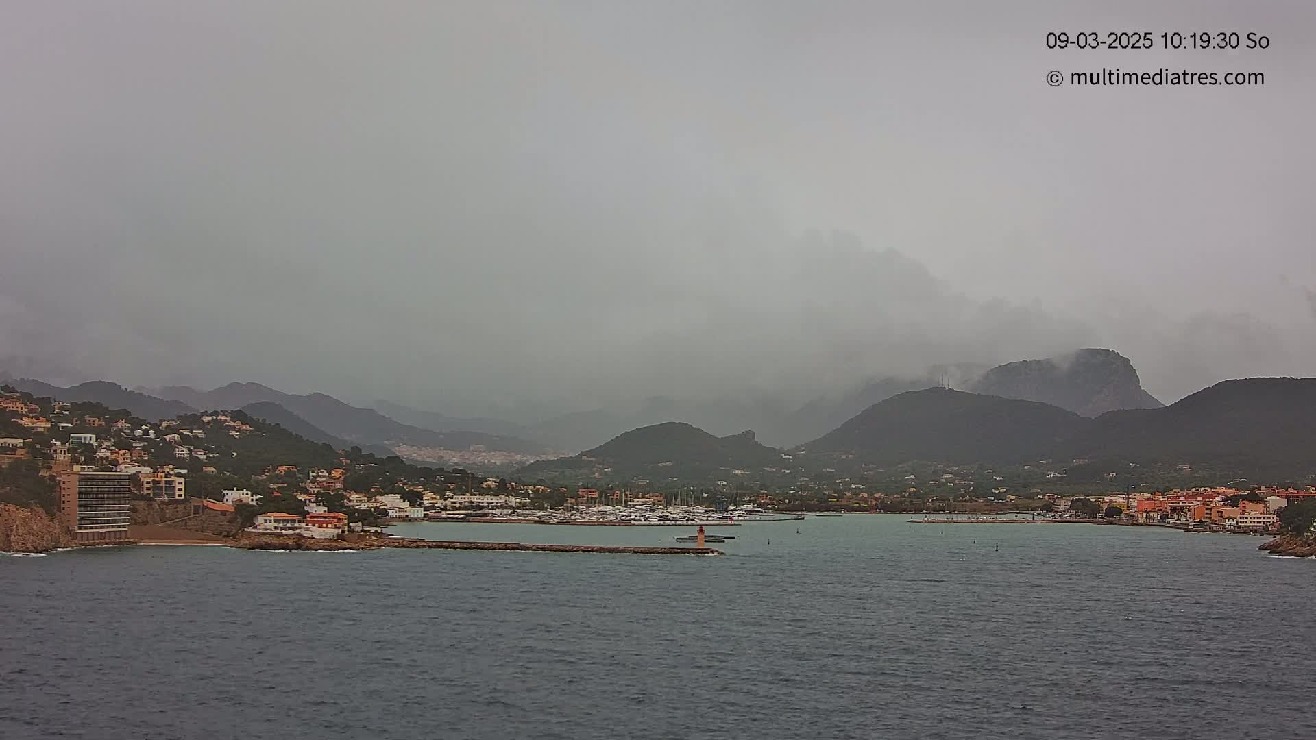 A coastal town nestled between mountains is shrouded in a hazy, overcast sky, with a calm body of water in the foreground.
