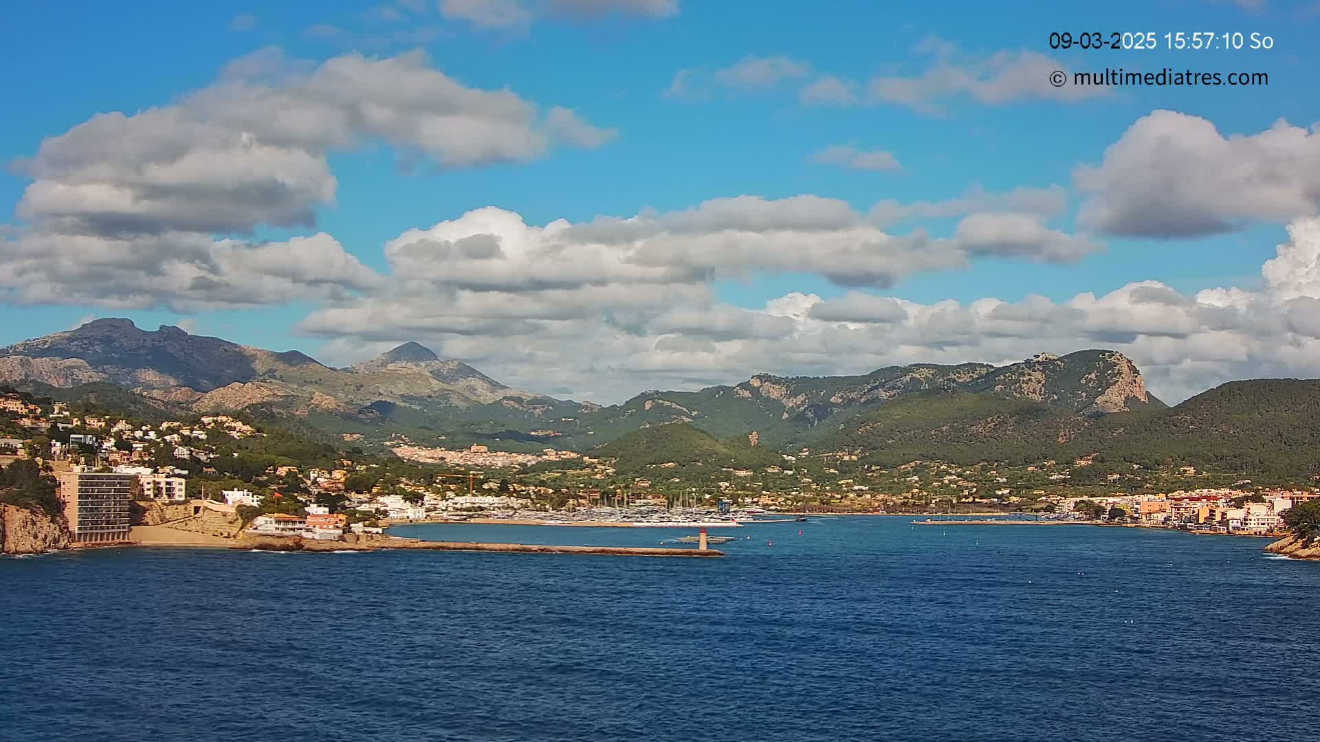 A coastal town nestled beneath mountains, with a marina and a partly cloudy sky above, is seen from across a body of water.