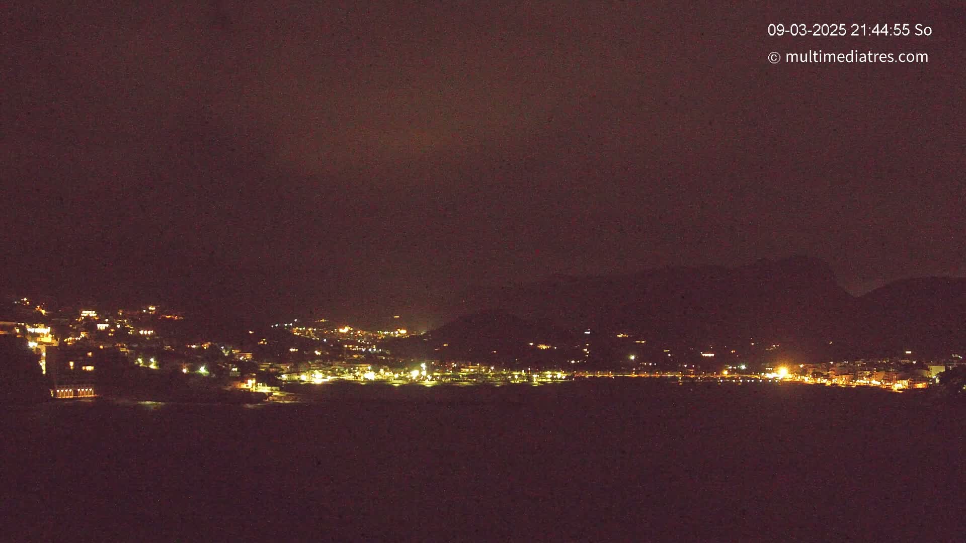 A nighttime long shot shows a coastal town illuminated with scattered lights, nestled against dark hills under a dark sky.