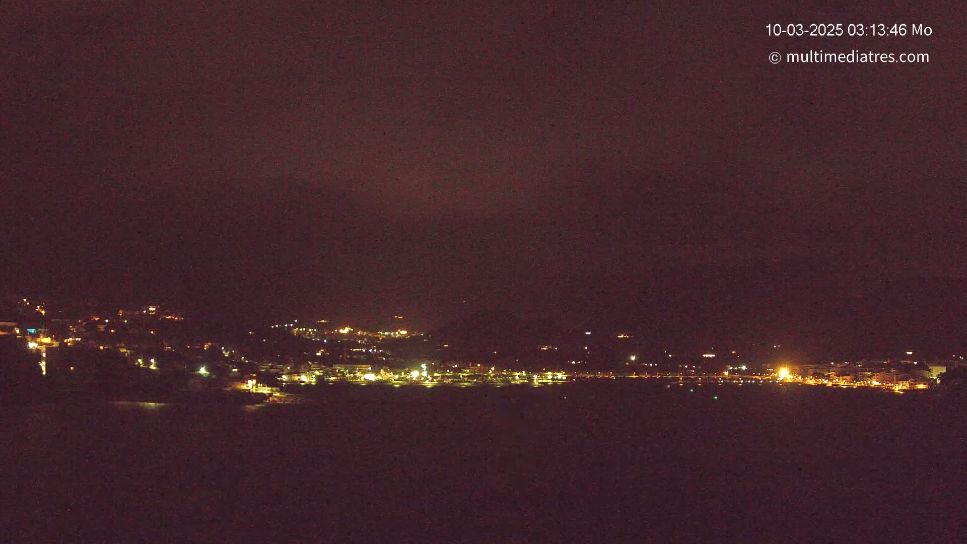 A nighttime long shot shows a coastal town's lights spread across a hillside and along a dark body of water under a dark sky.