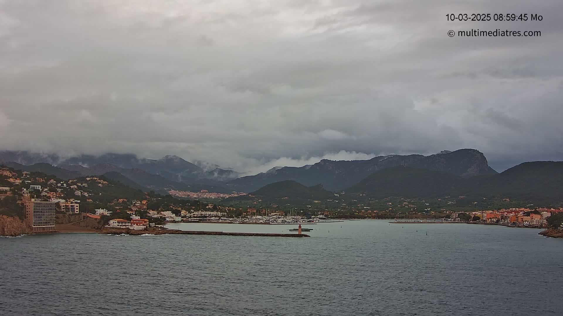 A coastal town nestled below overcast skies and mountains, features a harbor with numerous boats.