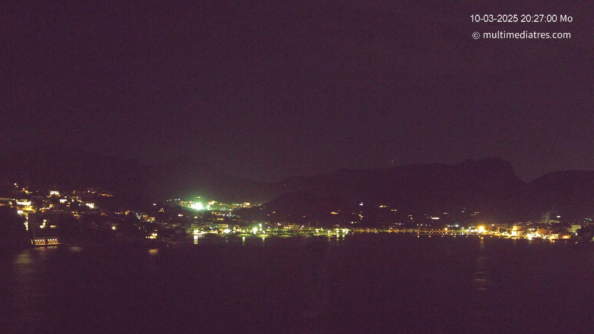 A nighttime view of a town nestled on a hillside next to a dark body of water, under a clear, dark sky.