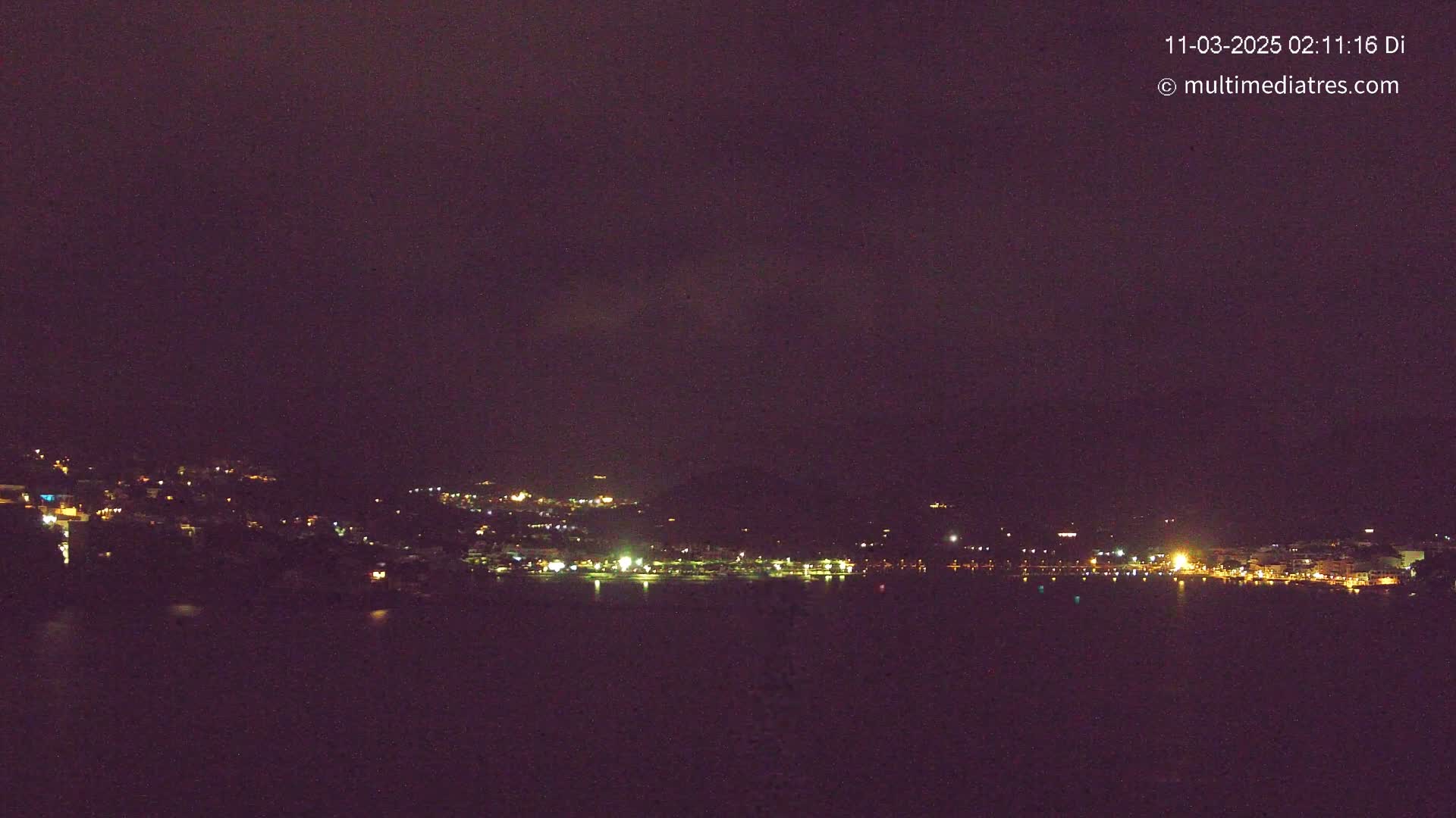 A nighttime view of a town situated on a body of dark water, under a dark, overcast sky.