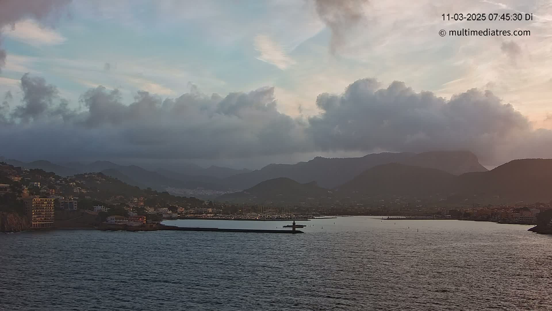 A coastal town nestled in mountains is viewed from across a calm body of water under a cloudy sky.
