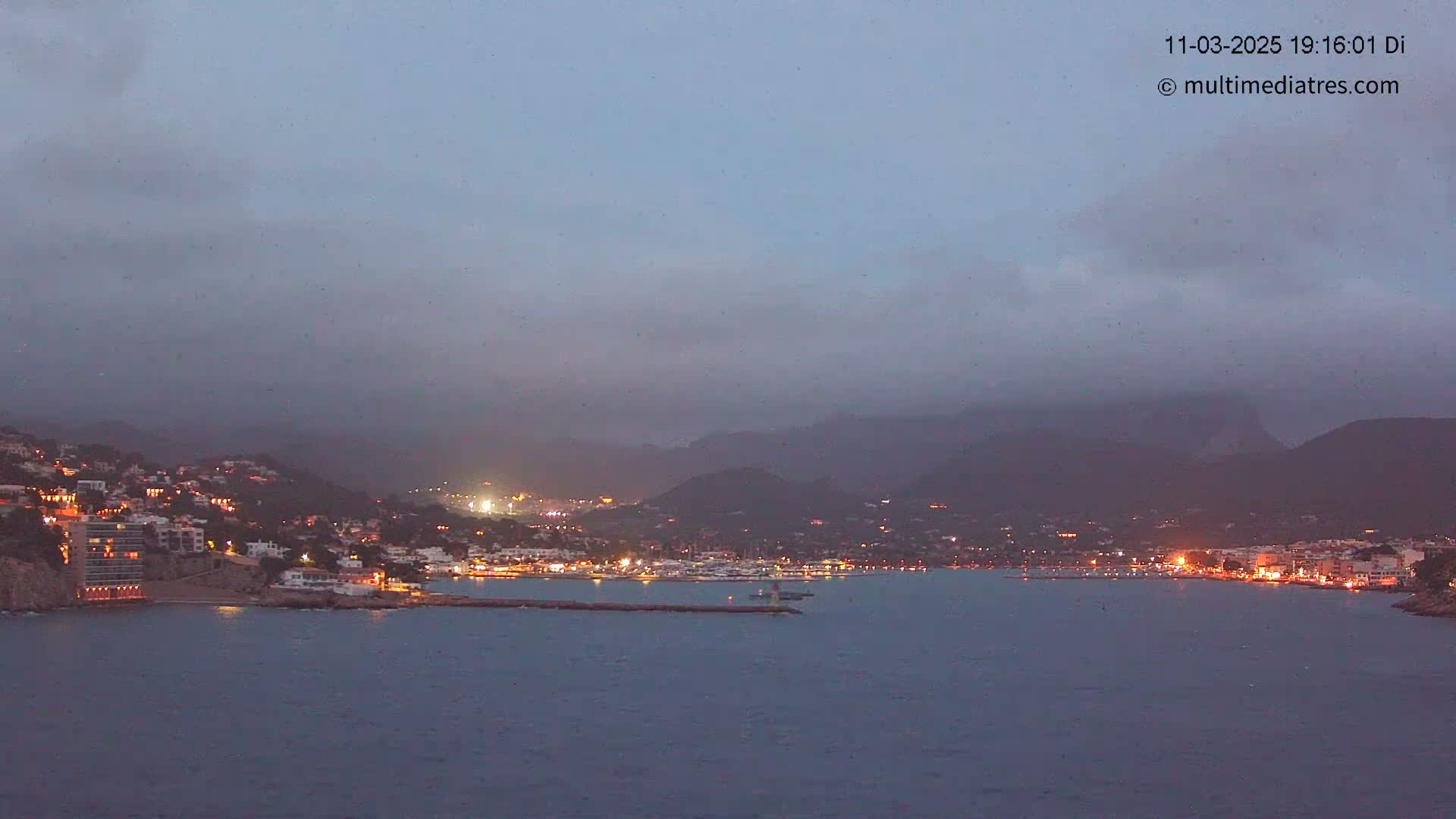 A nighttime coastal town with lights illuminating buildings nestled against dark mountains under a hazy, overcast sky.