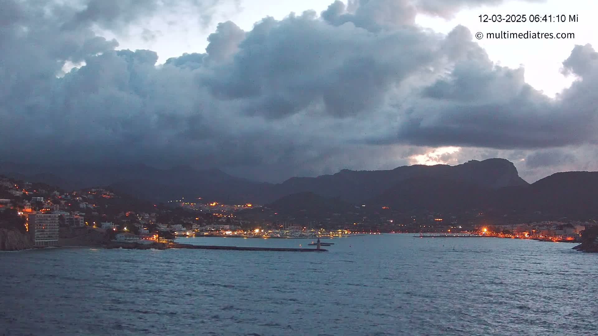 A coastal town at dusk is nestled beneath a dark, cloudy sky, with its lights twinkling against a dark mountain range and a calm, dark-blue sea.