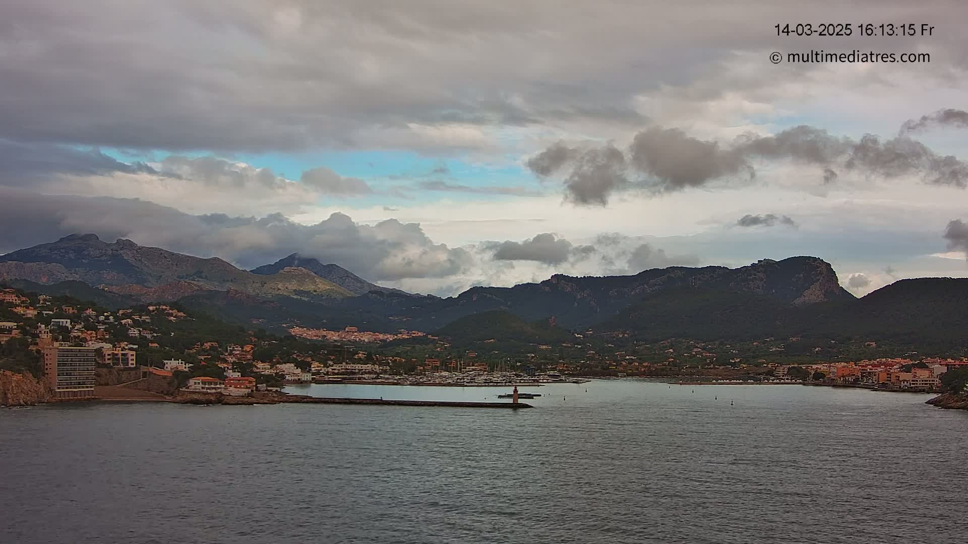A coastal town nestled between mountains under a cloudy sky, with a harbor filled with boats.
