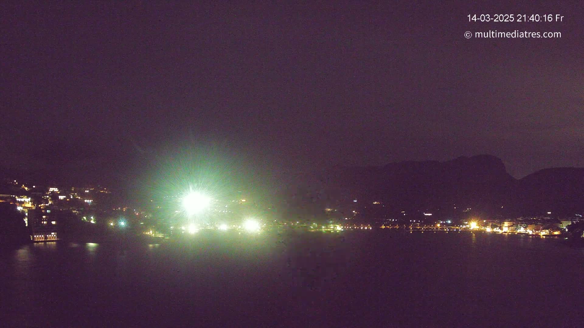 A nighttime long shot of a town illuminated along a dark body of water, backed by dark hills under a clear, dark sky.