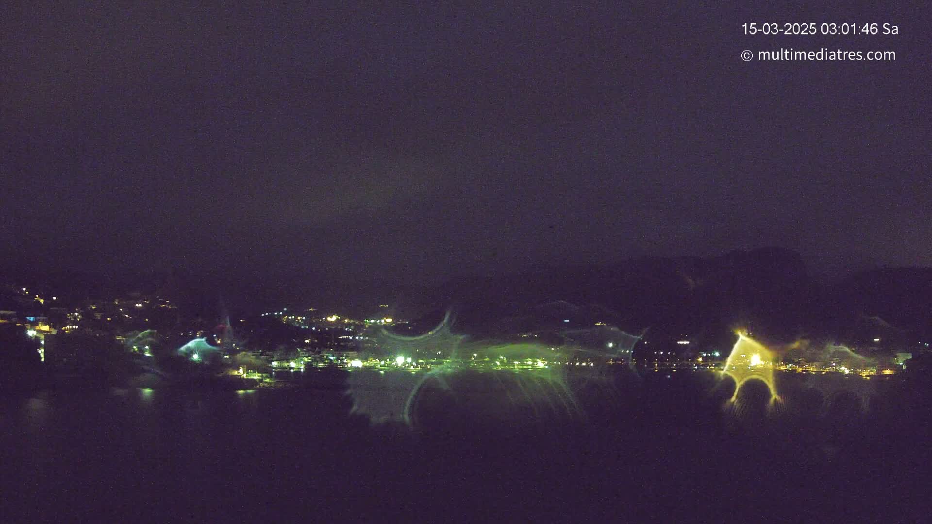 A nighttime long-range view of a city across a body of water, illuminated by lights and possibly exhibiting atmospheric distortion or lens effects.