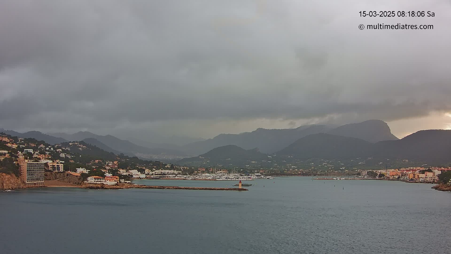 A coastal town nestled against dark mountains under an overcast sky, with a calm harbor filled with boats.