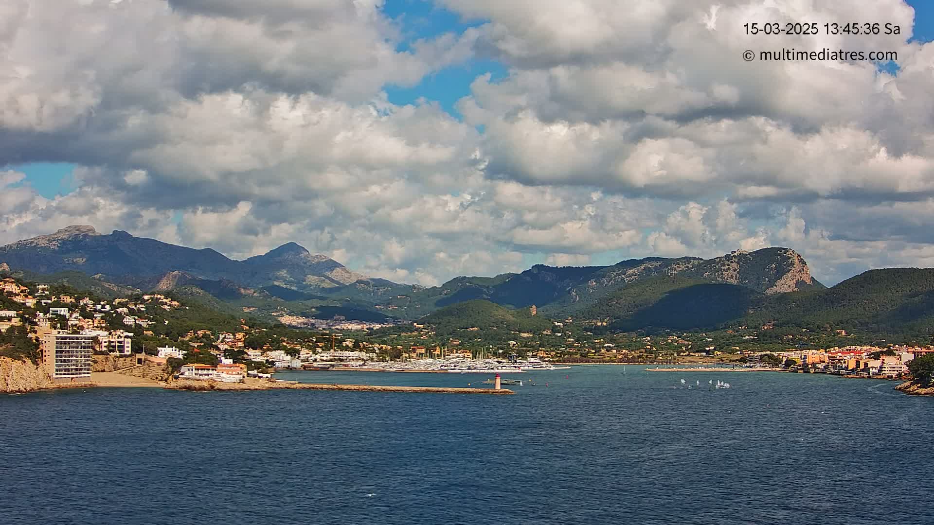 A partly cloudy day reveals a coastal town nestled between mountains and a calm bay filled with sailboats.