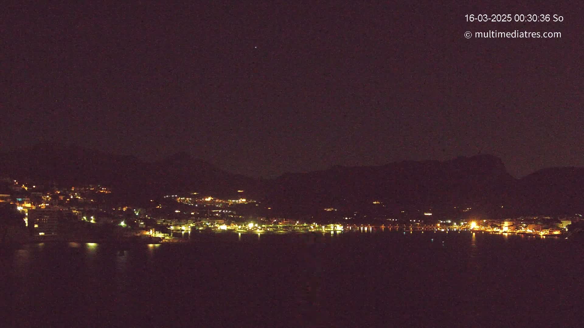 A nighttime view of a town nestled between dark hills and a calm body of water, illuminated by scattered lights.