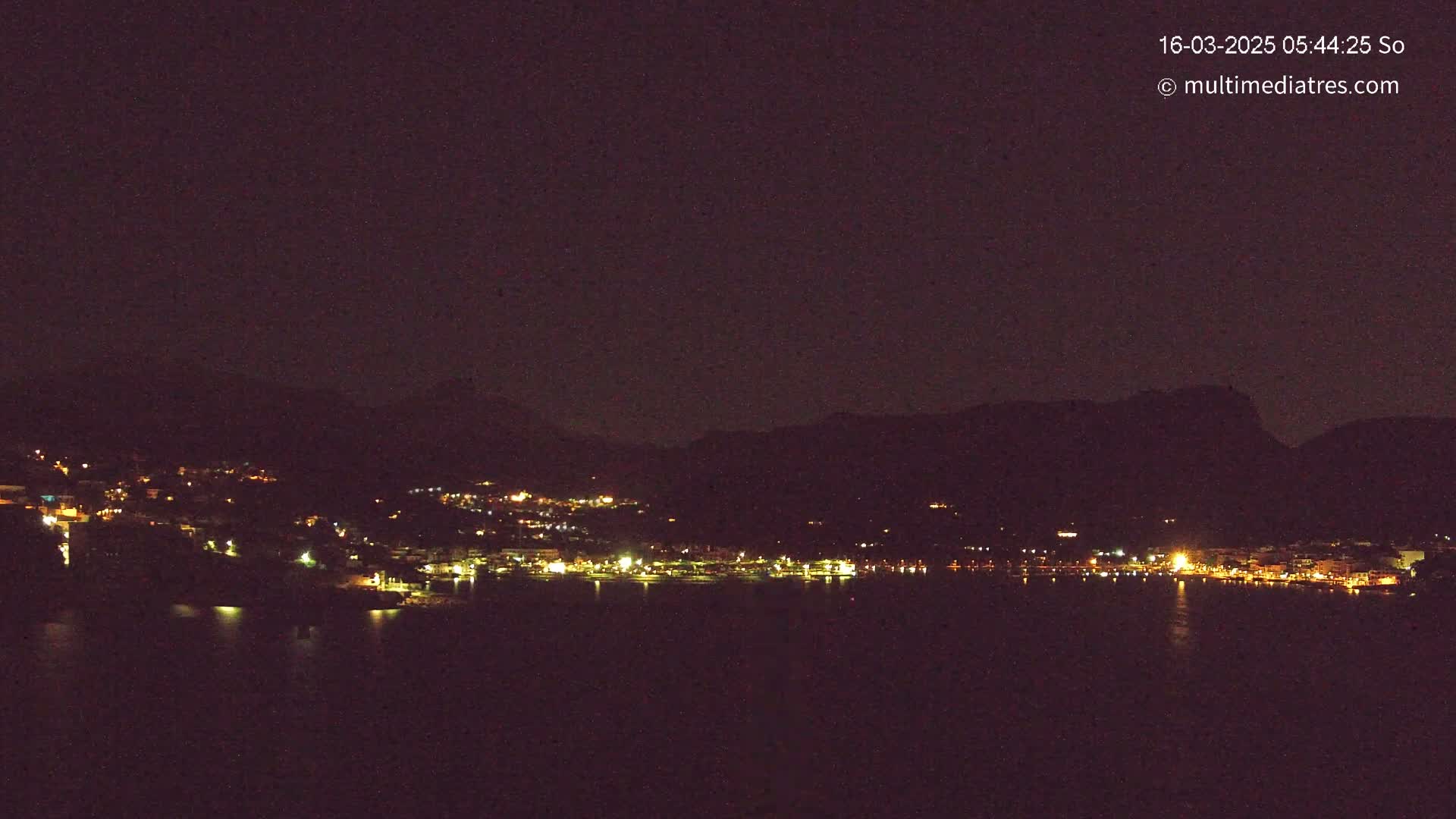 A nighttime view of a coastal town with lights illuminating buildings along the water, nestled against dark mountains under a clear, dark sky.
