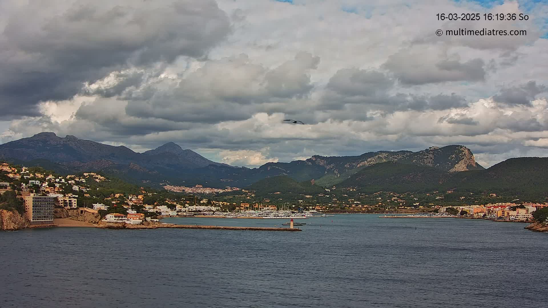 A coastal town nestled beneath a mountain range under a cloudy sky, with a harbor filled with boats.