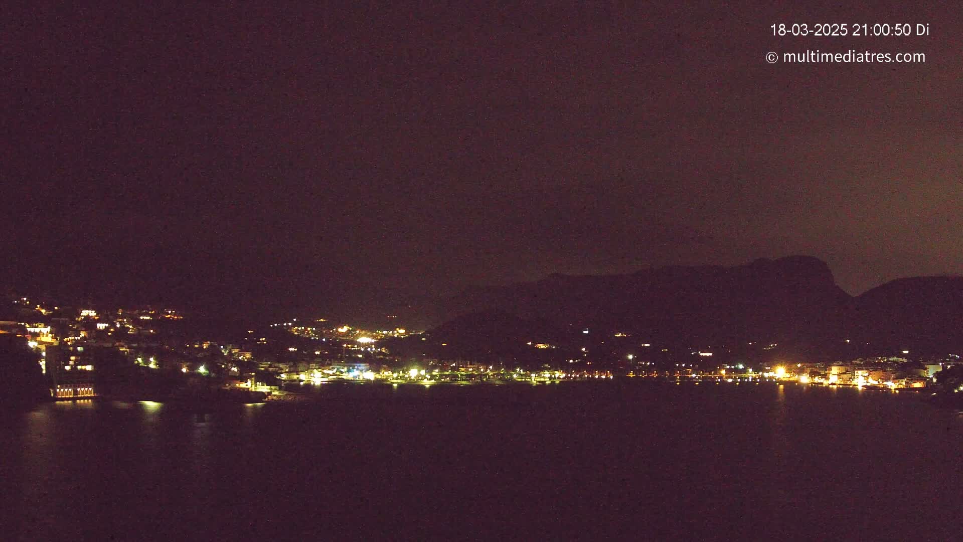 A nighttime long shot shows a coastal town illuminated with scattered lights, nestled against a dark mountain under a clear, dark sky.