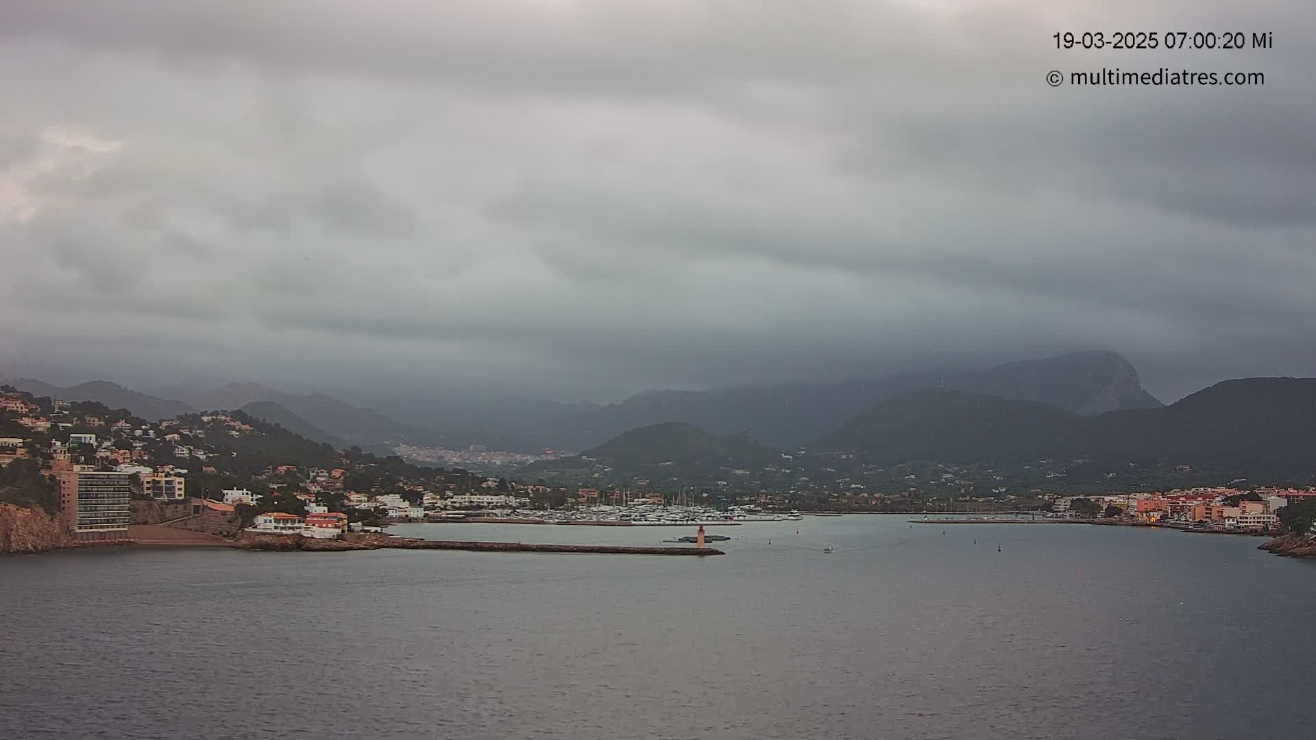 A coastal town nestled among mountains is viewed from across a calm body of water under an overcast sky.