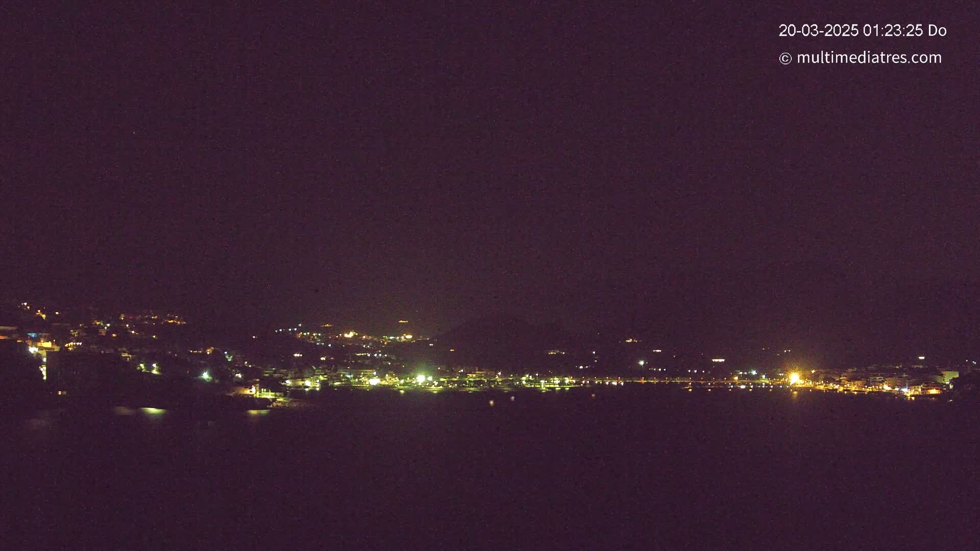 A nighttime long shot shows a coastal town illuminated with lights across a dark body of water under a clear, dark sky.