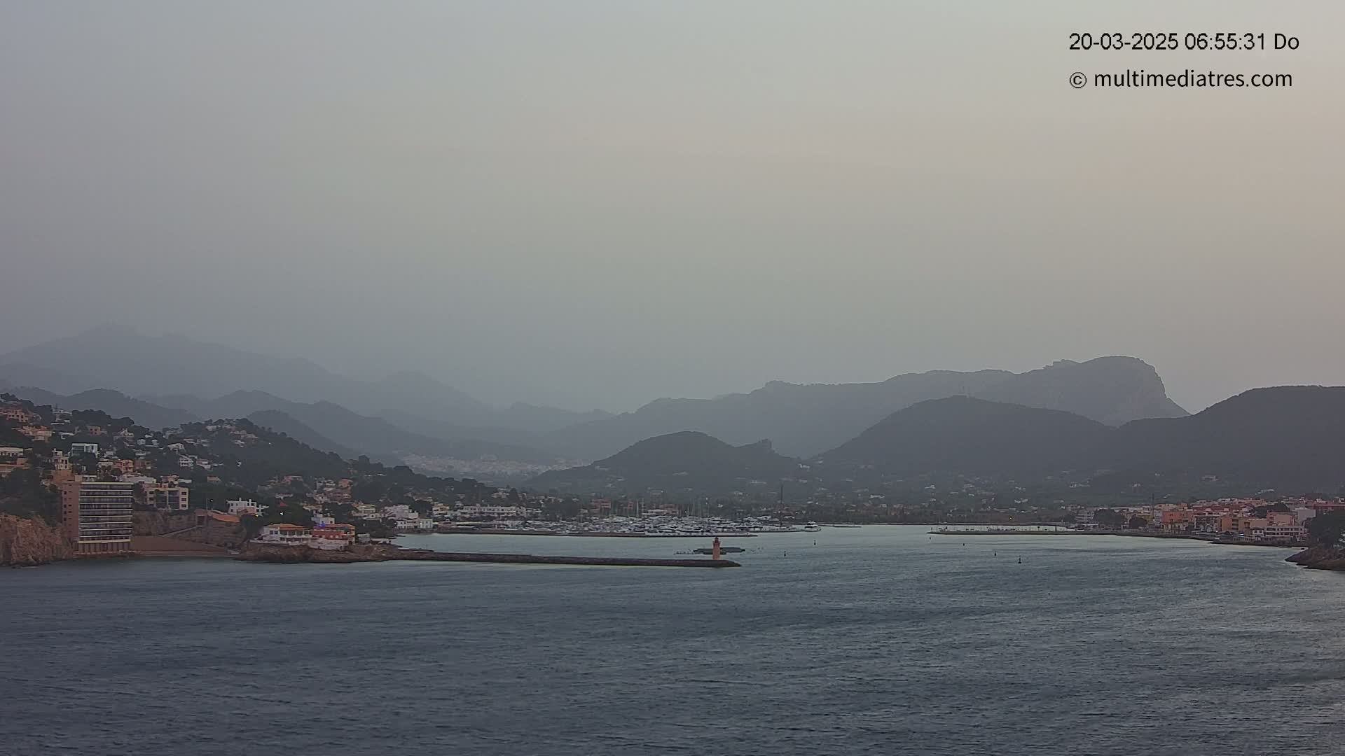 A hazy view shows a calm body of water with a marina and buildings along the coast, backed by distant mountains under a gray sky.