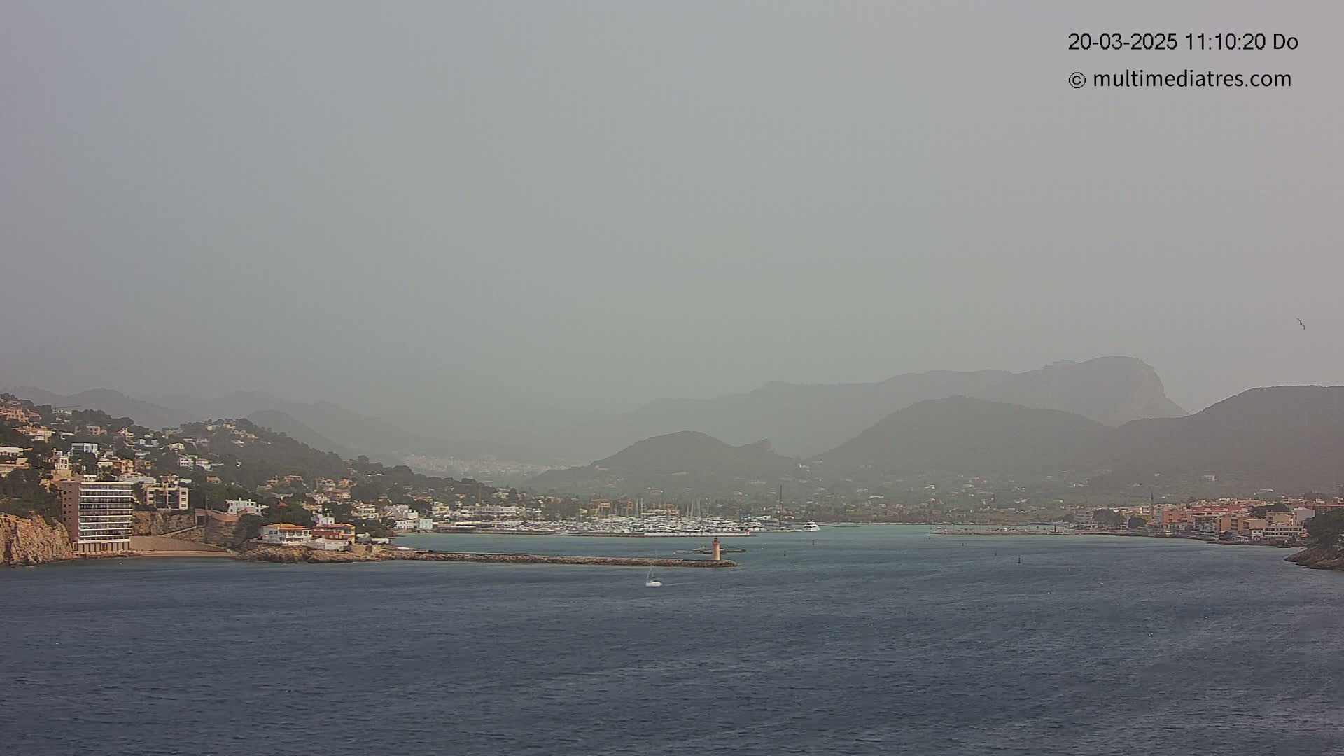 A hazy, dusty coastal scene shows a calm body of water with a marina and hillside buildings under a muted sky.