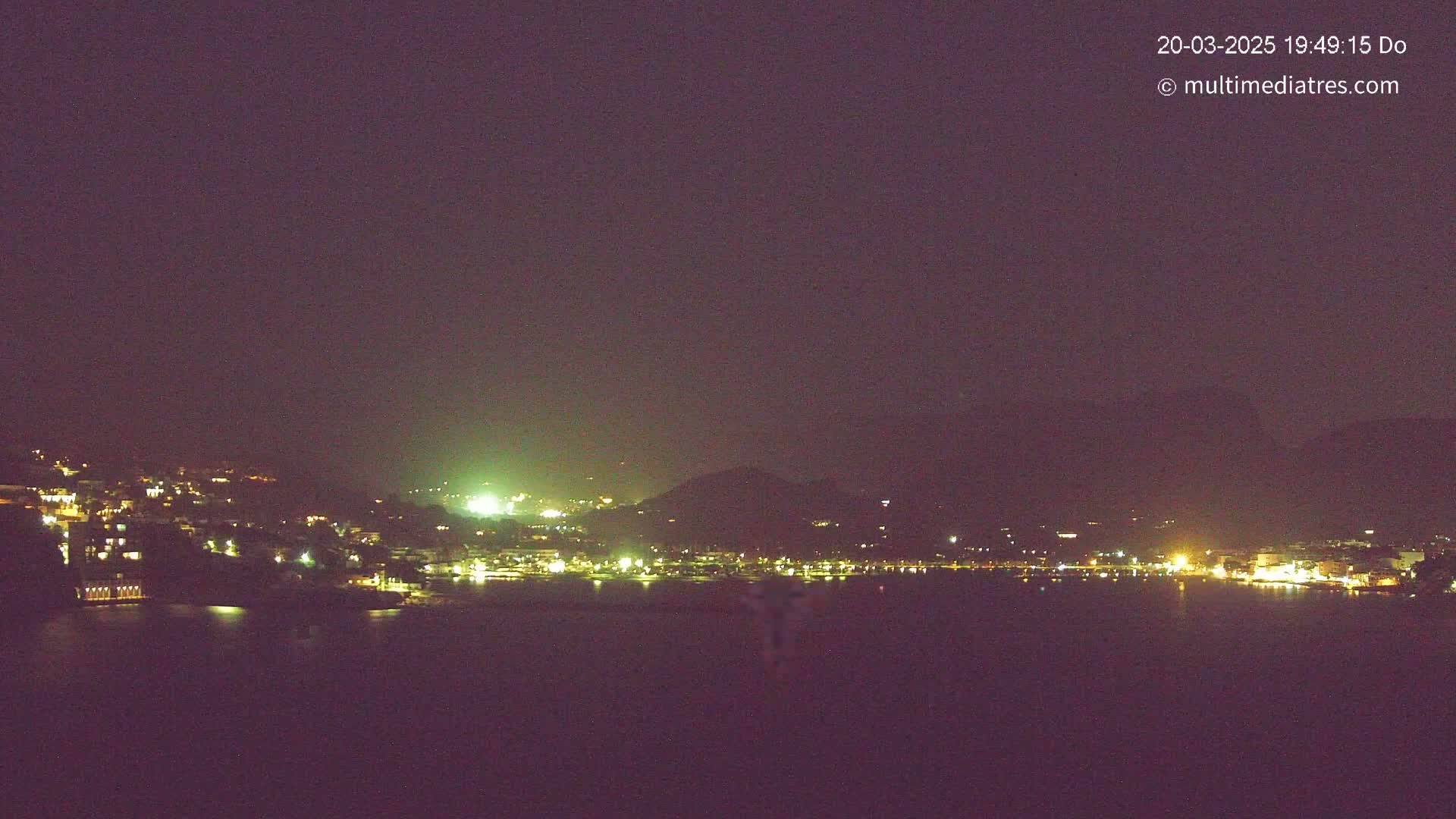 A nighttime, long-range view shows a town's lights spread across a hillside and reflected in a dark body of water under a clear, dark sky.
