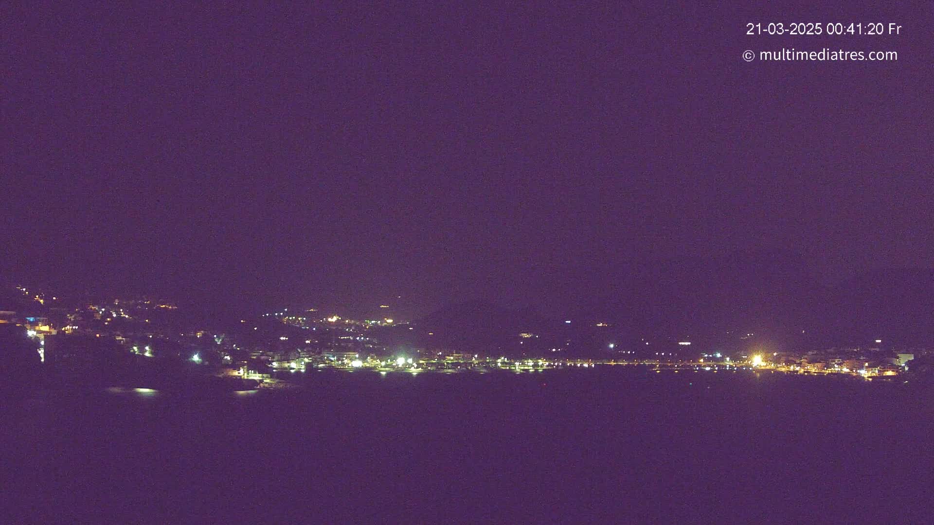 A dark night scene shows a coastal town with scattered lights along the shoreline and hillside, under a clear, dark purple sky.