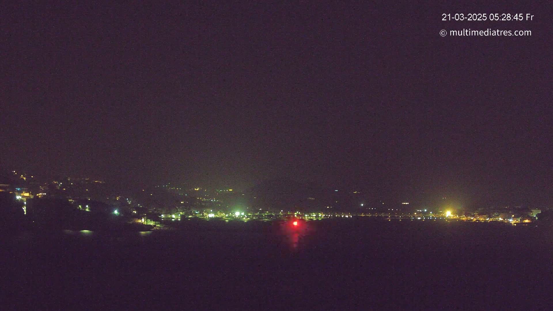 A nighttime long shot shows a coastal town's lights spread across a dark hillside, with a single red light visible in the water below.