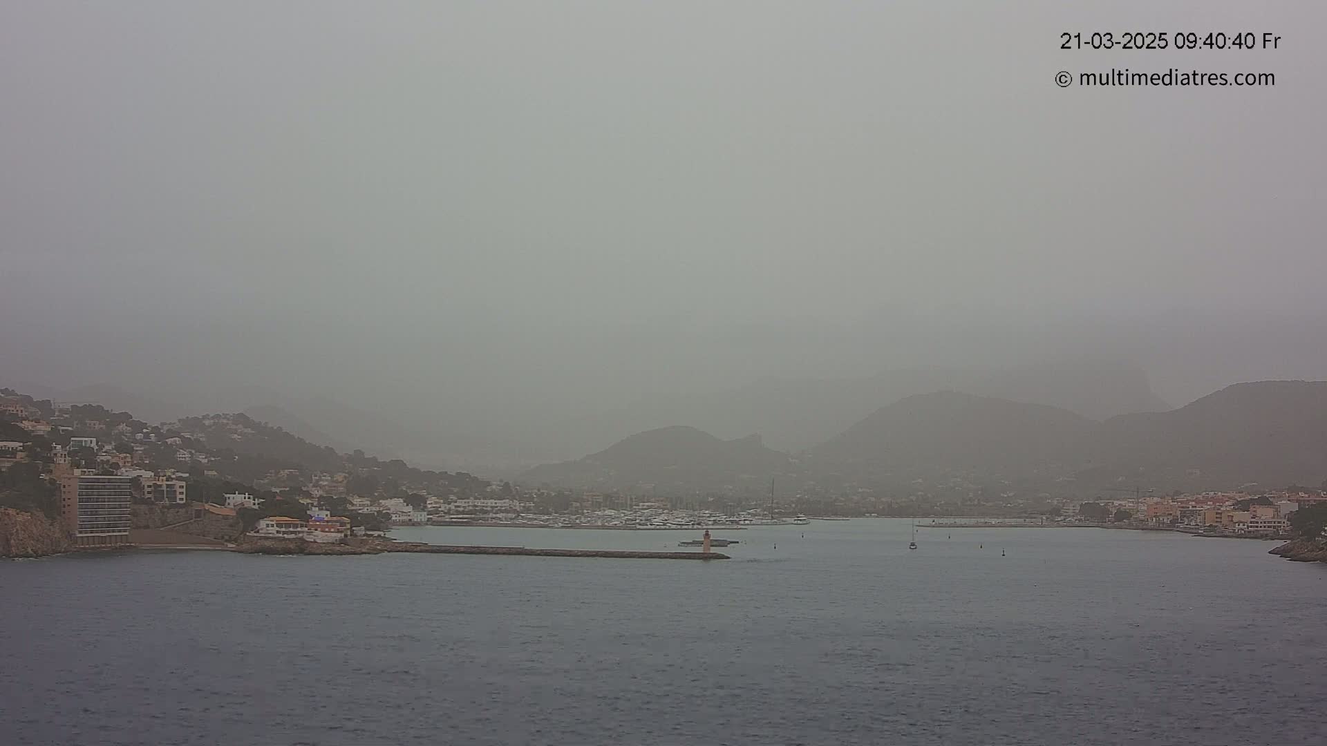 A hazy, dusty view of a coastal town nestled between hills and a calm body of water, with a small marina visible.